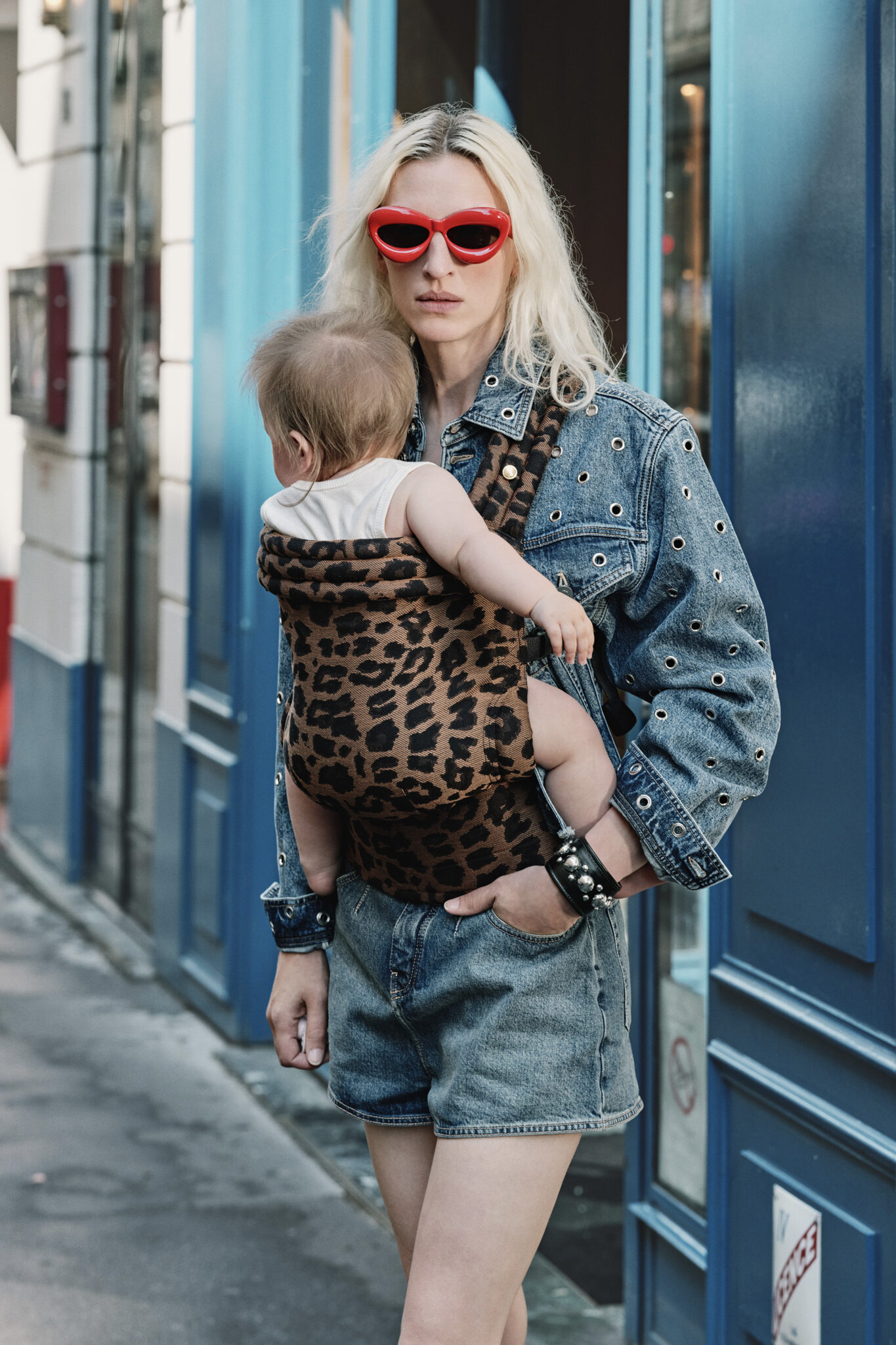 Fashion-forward parent wearing red cat-eye sunglasses and a denim jacket with grommet detailing, carrying a baby in a leopard print baby carrier. The duo exudes a chic urban vibe while strolling through a city street with blue accents in the background
