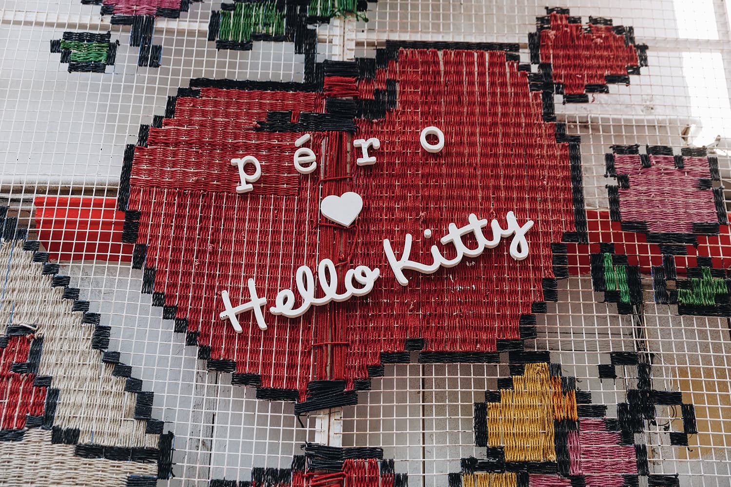 A woven art piece featuring a large red apple with the words 'péro ♥ Hello Kitty' in white letters, set against a grid background. Surrounding the apple are additional colorful woven shapes resembling flowers or fruit in shades of pink, green, and yellow, adding a playful, nostalgic feel to the design.