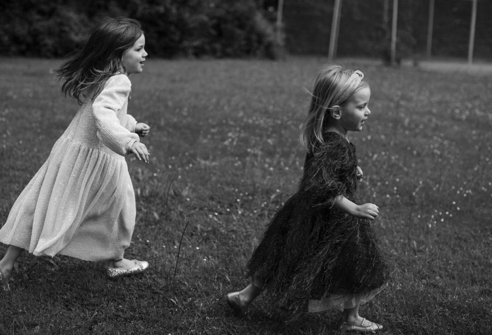 Two young girls are running across a grassy field in black and white, capturing their carefree movement. They both wear playful, long dresses that move with the breeze.