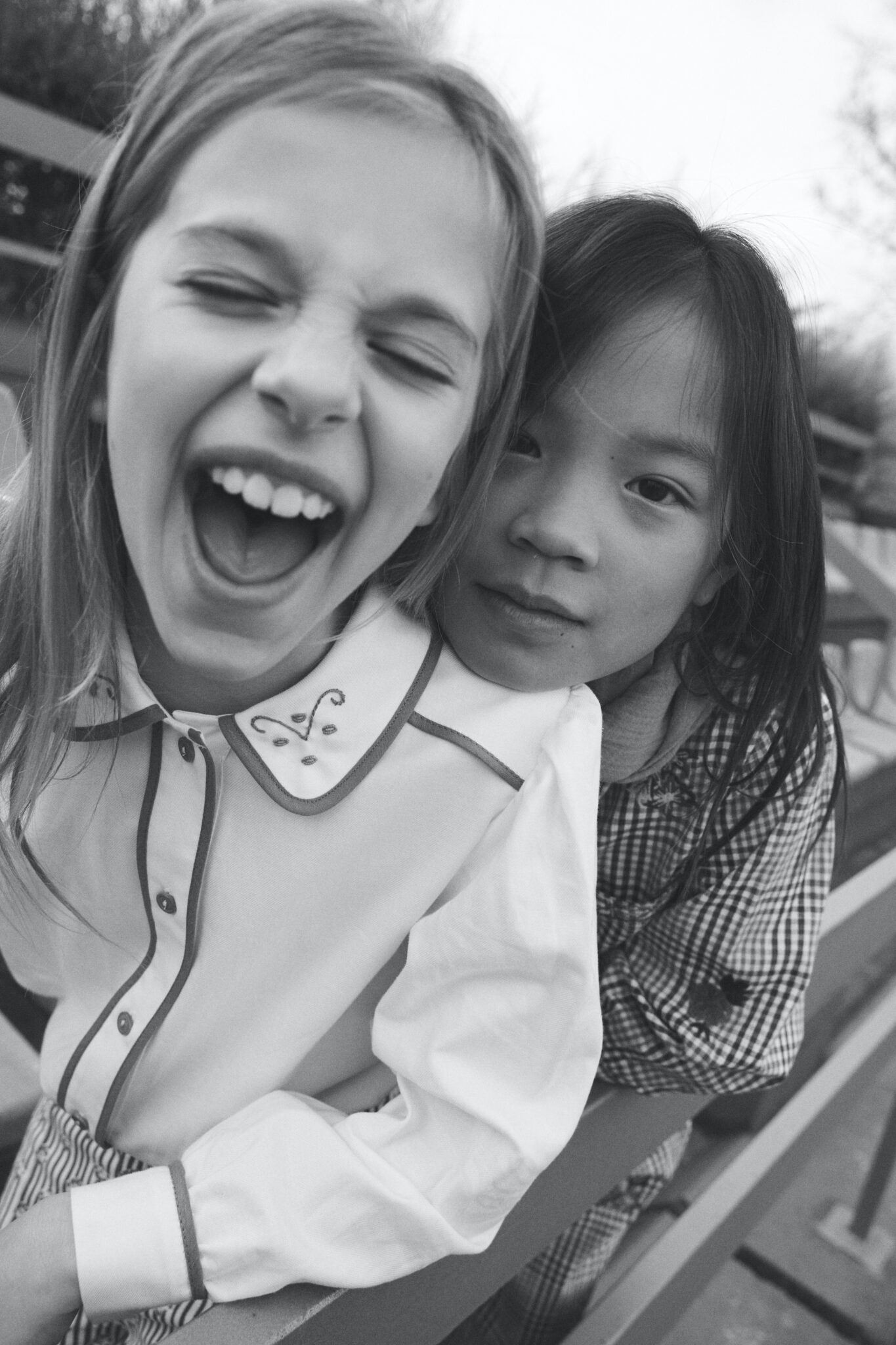 Two children smiling and posing together outdoors, showcasing Birinit's FW24/25 collection. The child on the left wears a light blouse with detailed embroidery on the collar, while the child on the right sports a gingham-patterned outfit. The photo is in black and white, emphasizing the playful and classic aesthetic of the collection.