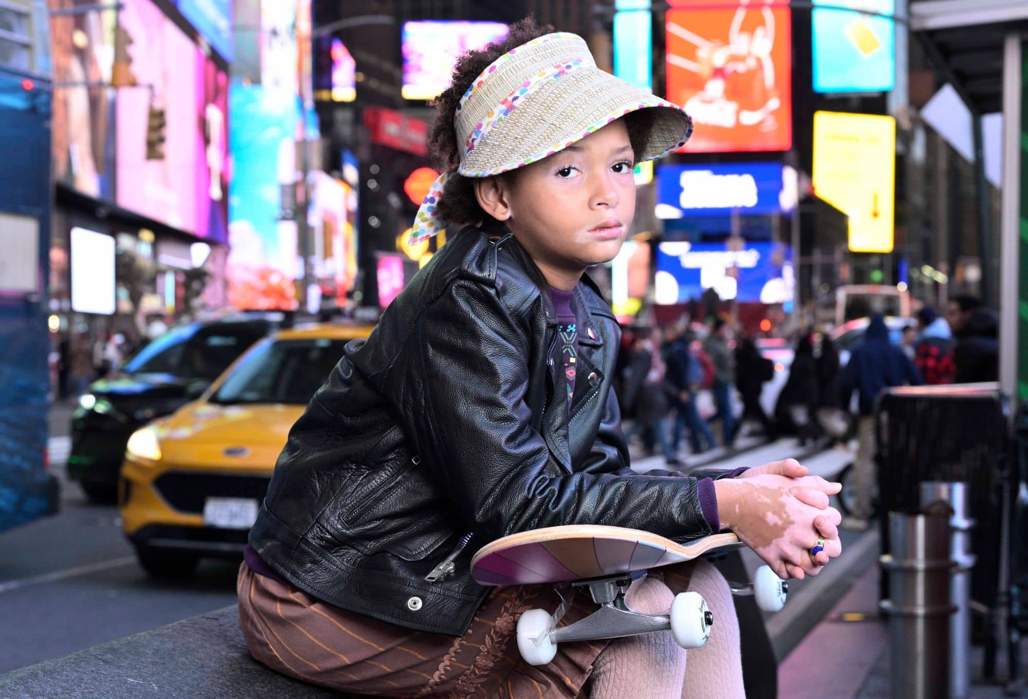 A young child sits on a ledge in Times Square, wearing a black leather jacket, patterned brown trousers, and a woven visor hat with colorful trim. They hold a skateboard and gaze thoughtfully into the distance, surrounded by the vibrant lights and bustling activity of the city. A yellow taxi is visible in the background, enhancing the urban atmosphere.