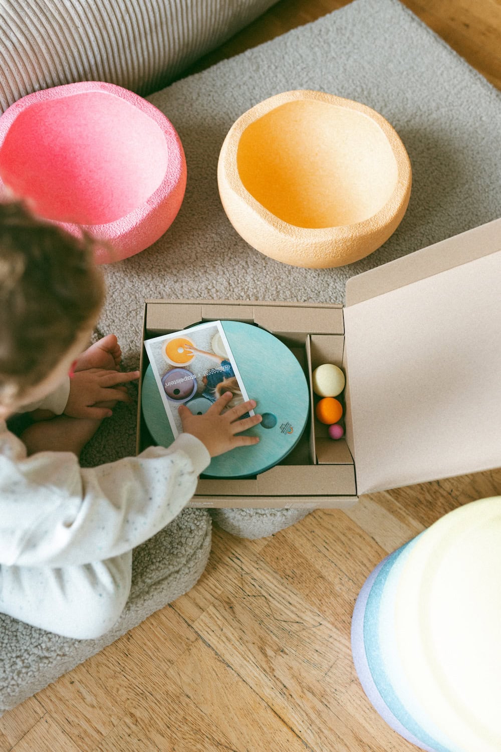 A young child in a cozy, neutral-toned outfit stands in a softly lit, minimalist living room, holding a round, pastel blue wooden disc in front of their face. In front of them, a stack of colorful, textured stacking bowls in pastel shades is carefully balanced on a cushioned platform, with a few additional wooden discs scattered nearby. The background features a beige corduroy sofa adorned with lavender and pink throw pillows, a white dome-shaped lamp, and a framed Matisse print resting against the wall. The scene exudes a calm, Scandinavian-inspired aesthetic with a playful, childlike touch.