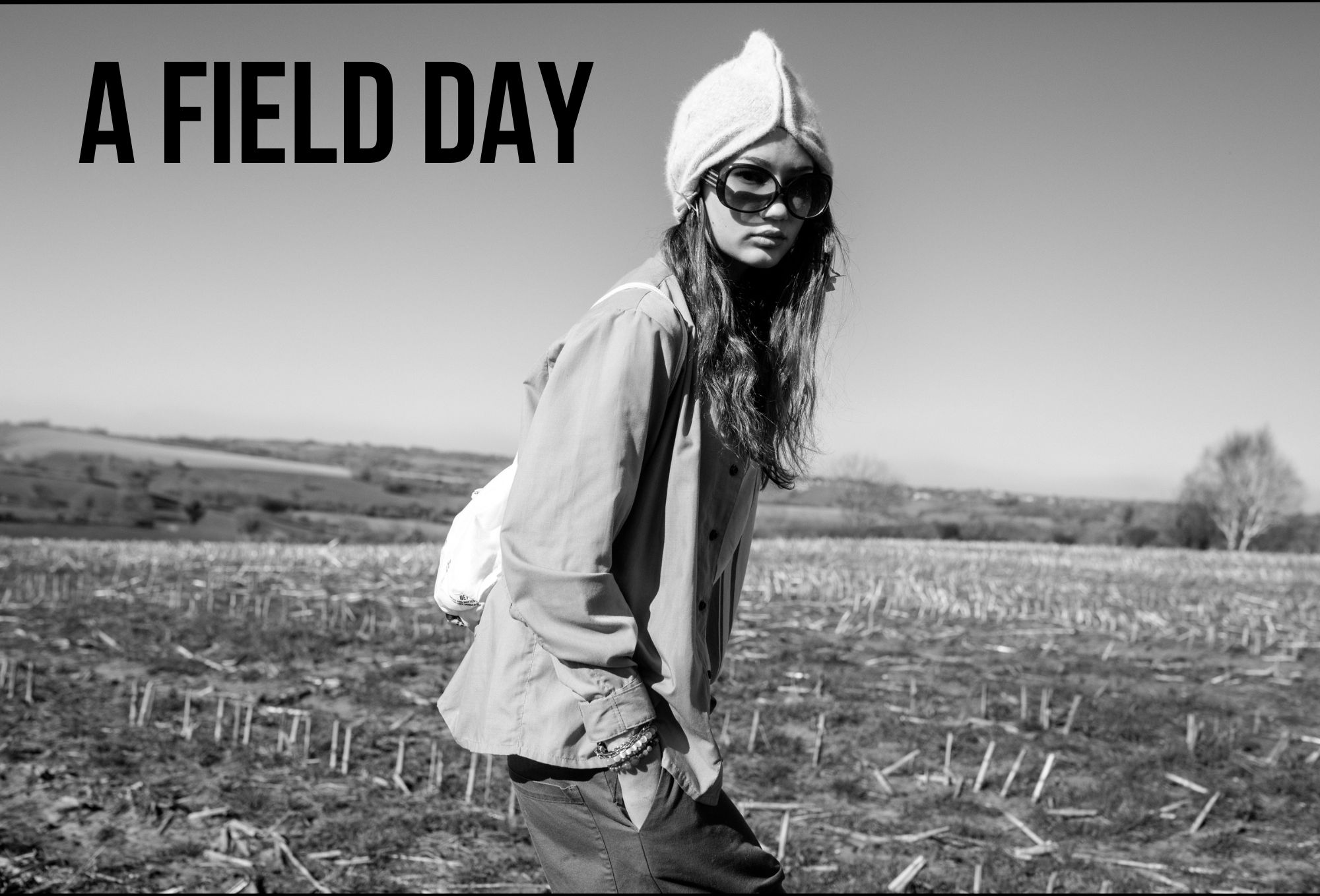Girl in a field wearing oversized sunglasses, a pointed wool hat, and a loose button-up shirt, with a backpack on her shoulder. The photo is black and white with countryside in the background. Large text reads