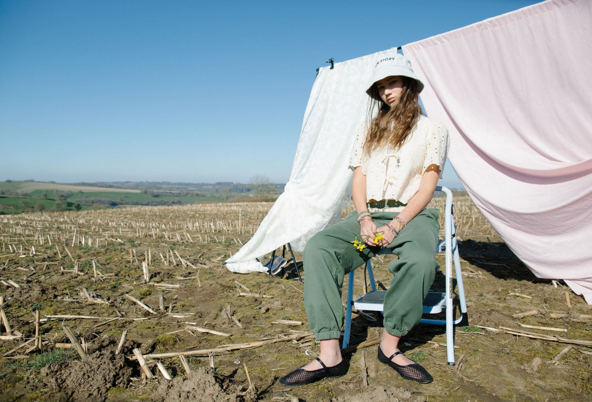 Girl sitting on a folding chair in a harvested field, wearing a white eyelet blouse, green jogger-style trousers, black mesh flats, and a white bucket hat with text. Behind her hang pink and white sheets, creating a soft makeshift backdrop. She holds small yellow flowers in her hands, with bracelets adorning both wrists. The sky is clear and blue.