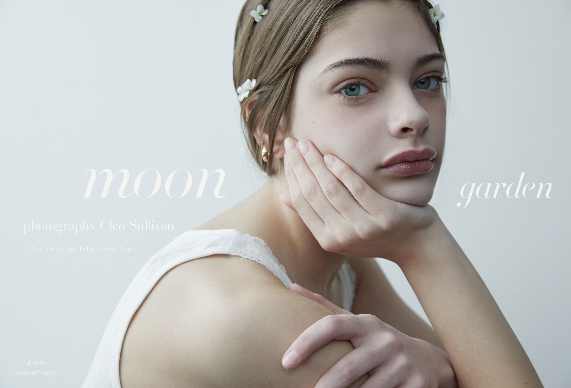 Ritratto ravvicinato di giovane modella con occhi azzurri, mano sul viso, top bianco in pizzo e fermagli a fiore tra i capelli; editoriale moda “Moon Garden” di Cleo Sullivan.