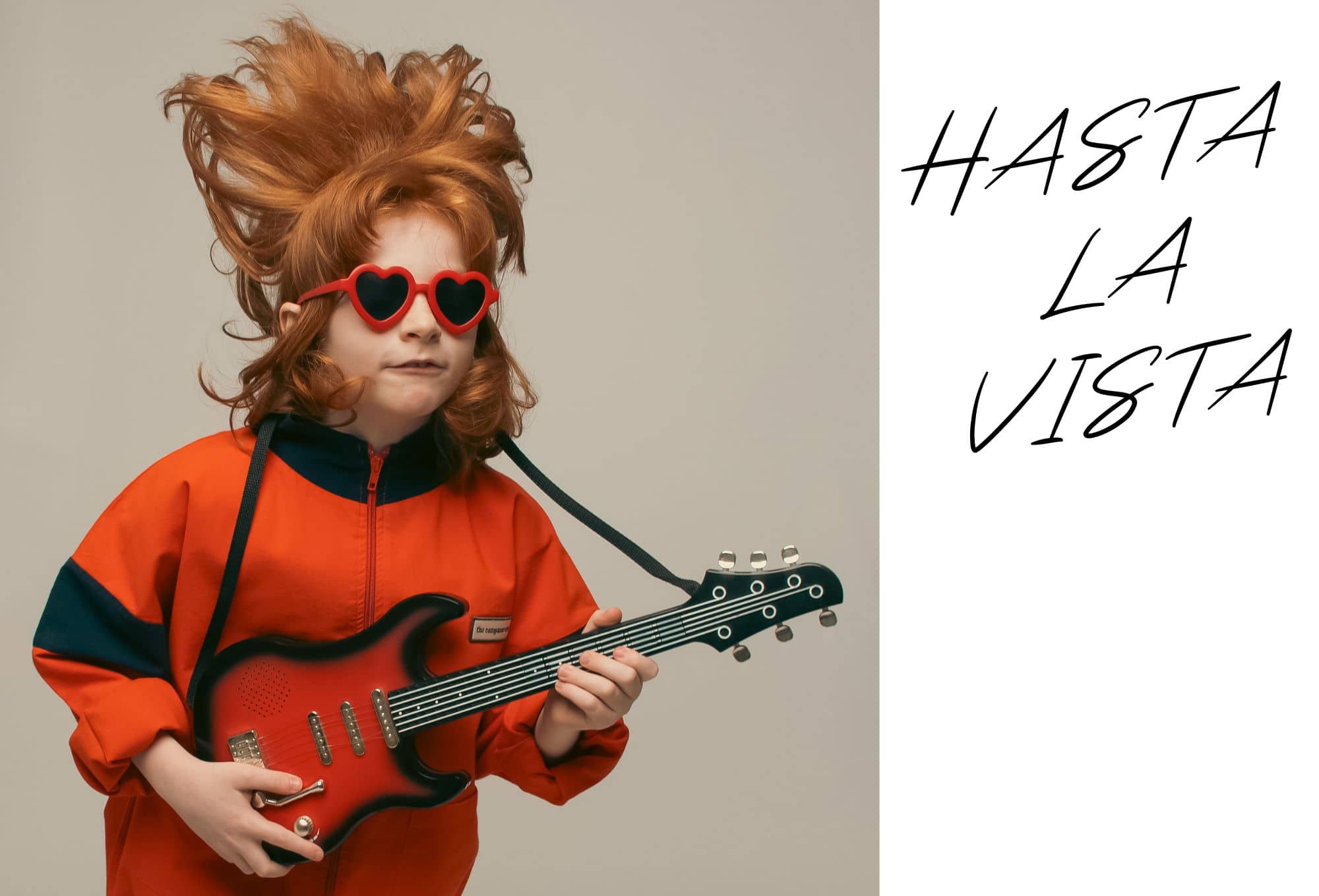 A red-haired child with voluminous hair and heart-shaped sunglasses rocks out with a toy guitar, wearing a bold orange tracksuit.