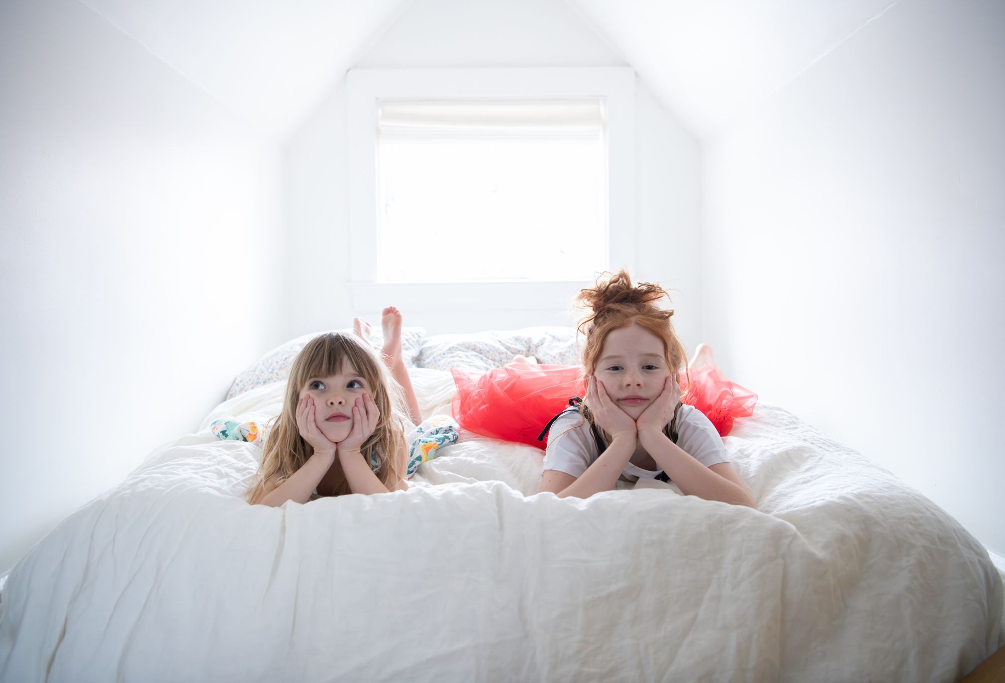 Two young girls lie on their stomachs atop a fluffy white bed in a bright, cozy attic room with white walls and a small window behind them. Both rest their faces in their hands with thoughtful or dreamy expressions. The girl on the left wears a floral outfit, while the girl on the right wears a red tulle dress over a T-shirt. Their bare feet are visible in the background, adding a carefree, relaxed vibe. Photo by Sabrina Helas.