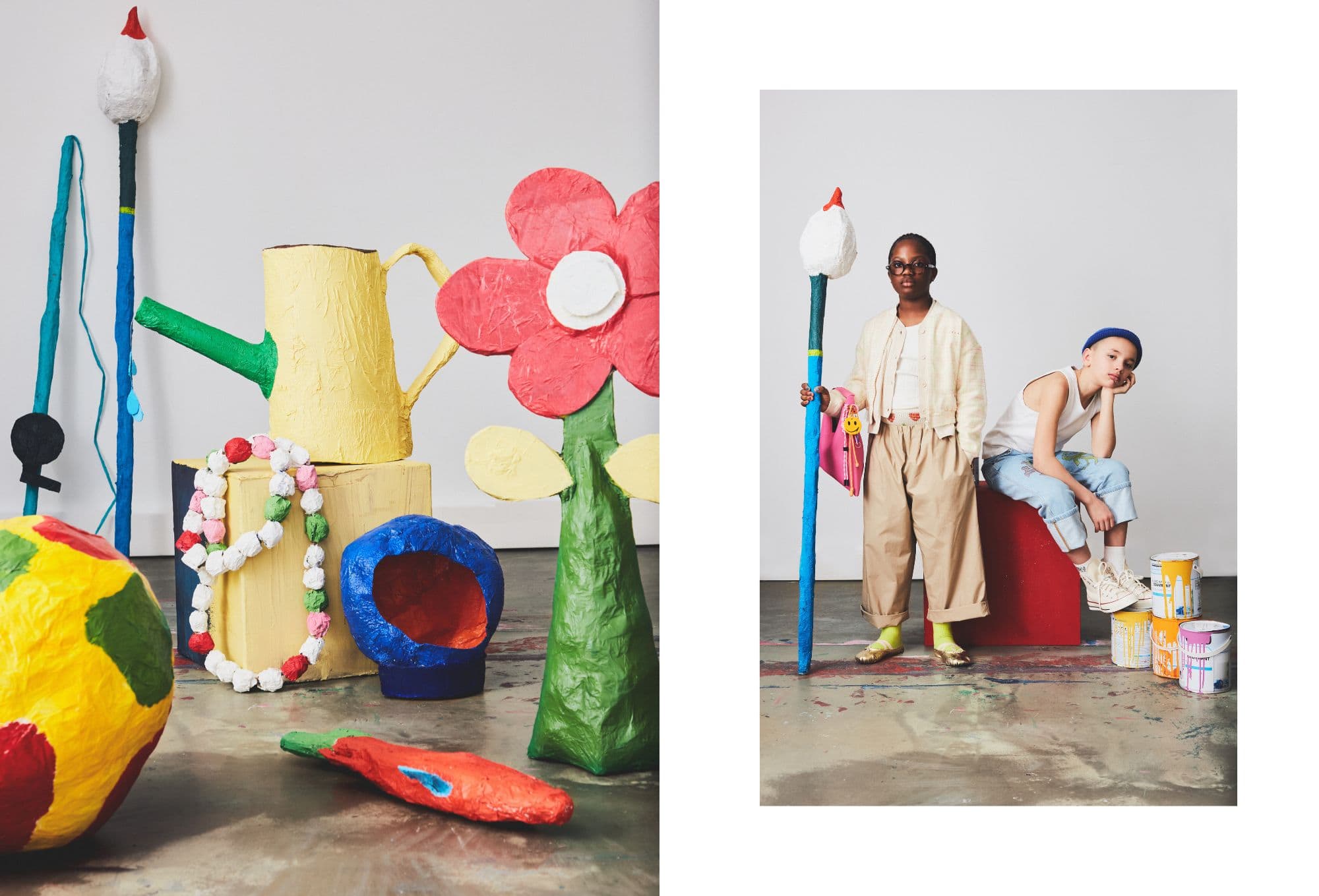Children’s fashion editorial with a playful, artsy vibe. On the left, a colorful still life features oversized paper-mâché props like a yellow watering can, red and pink flowers, a rainbow-colored globe, and chunky beads, all set on a concrete floor. On the right, two kids pose with confidence—one wears wide tan trousers, a cream jacket, golden socks with sandals, and holds a tall blue stick topped with a paper flower. The other sits on a red block, wearing rolled-up light denim pants, a white tank top, cream high-top sneakers, and a cobalt blue beanie, next to stacked paint cans splattered with bright colors.