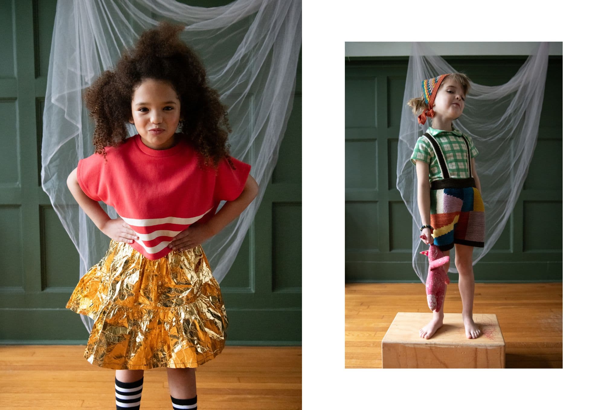 Two children pose confidently in a whimsical, indoor setting with a green paneled wall and sheer white drapery in the background. On the left, a girl with voluminous curly hair wears a bold red top with white stripes and a shiny, gold metallic skirt, paired with black-and-white striped socks. On the right, a child stands barefoot on a wooden box, wearing a colorful patchwork knit outfit with a green checkered top and holding a pink stuffed bunny. A crocheted headscarf completes the playful look. Photo by Sabrina Helas.