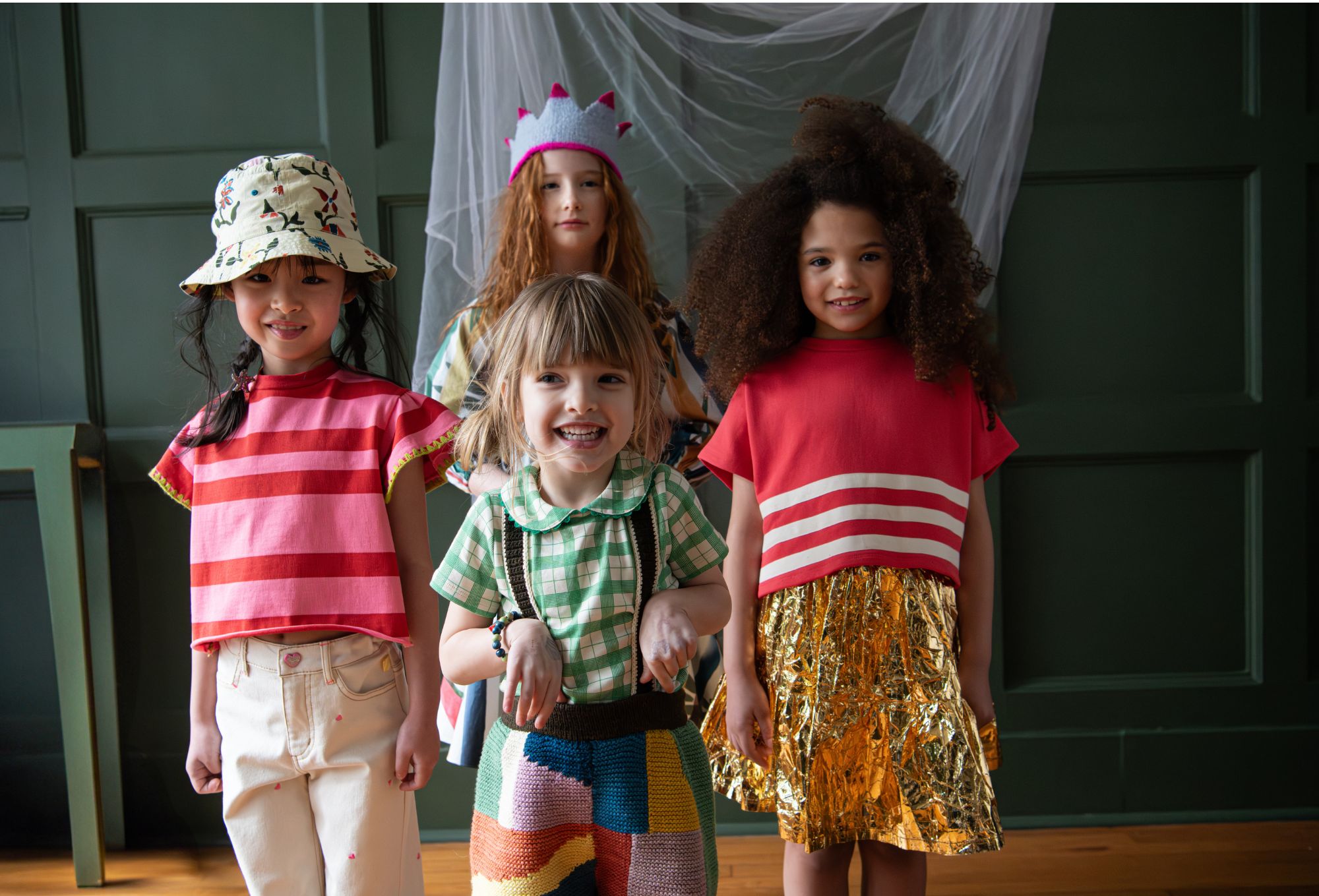 Alt text: Four children stand smiling in a playful indoor setting with a dark green paneled wall and draped white tulle in the background. The child in front wears a green checkered top with a colorful knit romper. To the left, a girl wears a pink striped top, cream jeans with tiny pink hearts, and a printed bucket hat. On the right, another girl wears a bold red top with white stripes and a shiny gold crinkled skirt. Behind them, a girl with long red hair and a felt crown peeks through, dressed in a multicolored striped dress. Photo by Sabrina Helas.