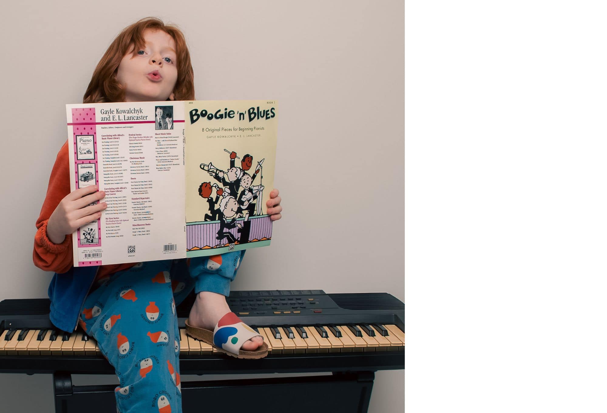 A child sitting on an electric keyboard, playfully singing while holding up a 'Boogie n’ Blues' piano book, wearing bright pajamas and mismatched sliders.
