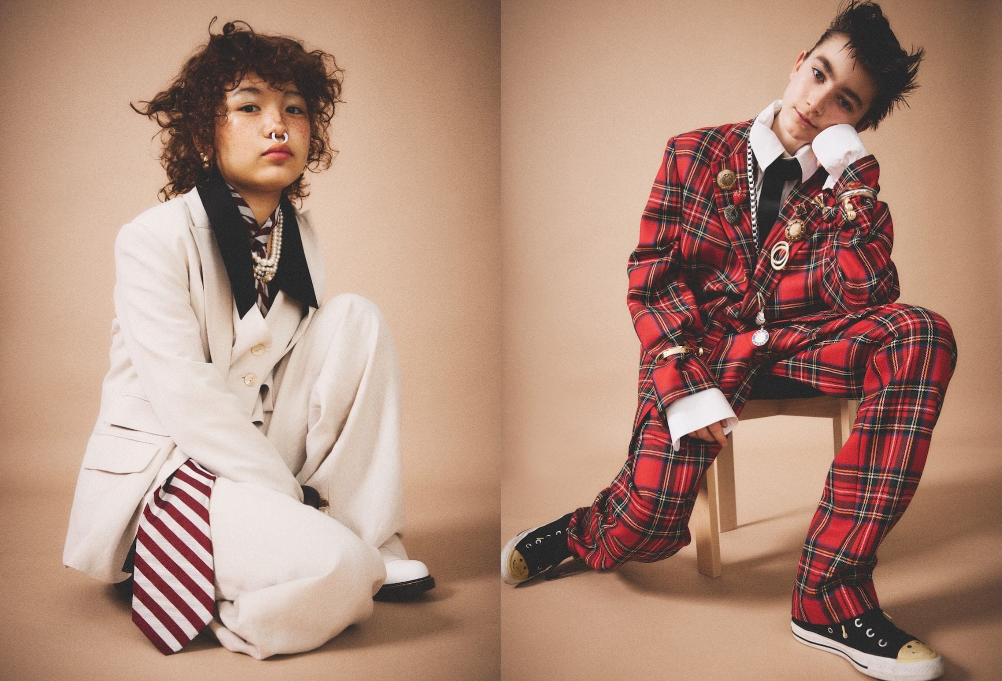 Fashion editorial by Molly Magnuson featuring two young models styled in exaggerated suiting. On the left, a model wears an oversized cream suit with a wide black collar, layered over a striped tie and pearl necklace, paired with a septum ring and curly hair. On the right, a second model sports a vibrant red tartan suit adorned with eclectic pins and chains, layered with a white shirt and black tie, and styled with black-and-white Converse sneakers. Both are posed against a soft beige background, blending punk, prep, and vintage influences with playful elegance.