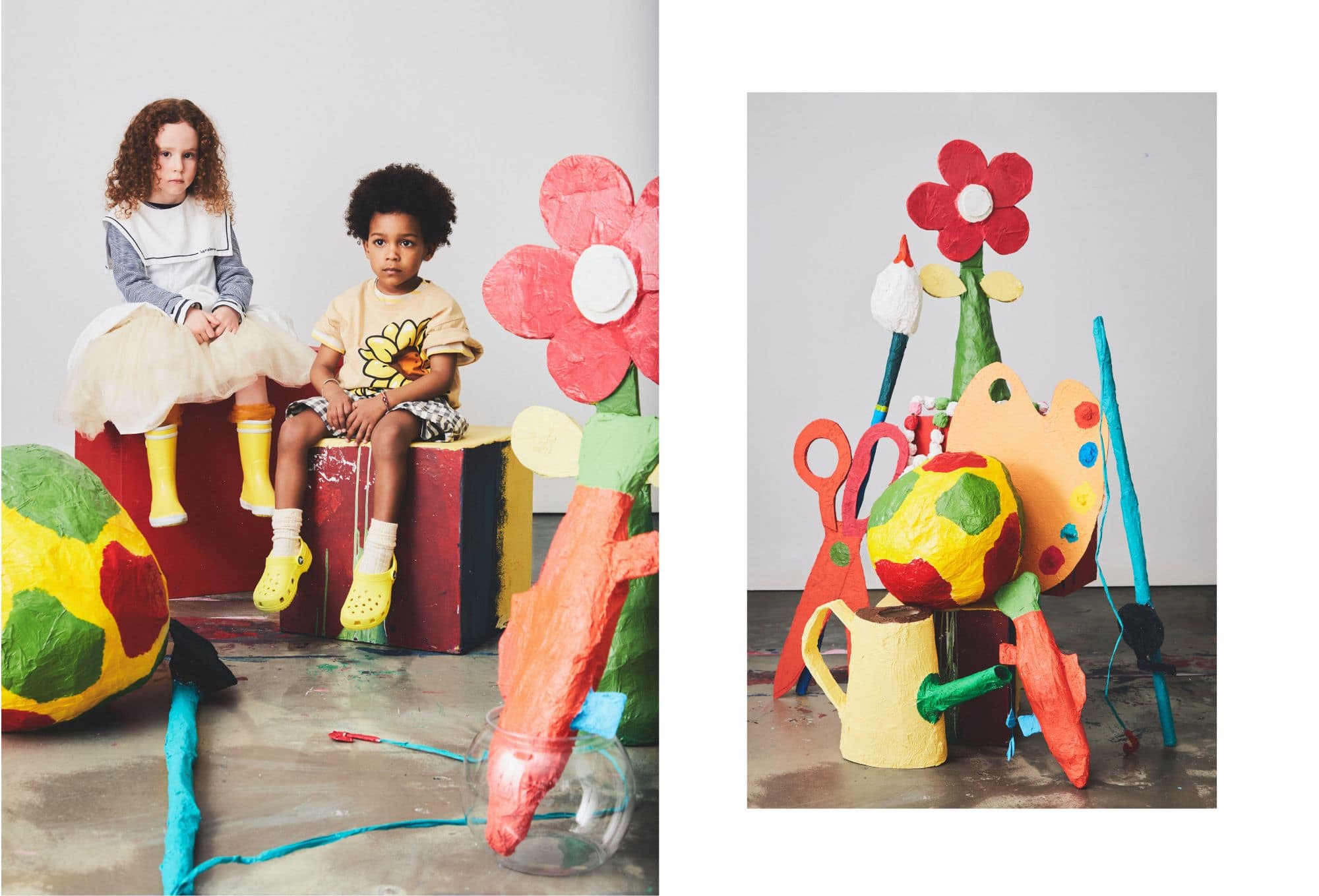 Colorful kids’ fashion editorial set in a handmade, oversized art studio world. On the left, two children sit on painted blocks surrounded by large paper-mâché props. The girl wears a sailor-style top with a cream tulle skirt and yellow rain boots, while the boy sports a beige T-shirt with a large flower print, checkered shorts, and yellow clogs with socks. On the right, an imaginative still life features giant paper-mâché scissors, paintbrushes, a palette, and a watering can, all painted in bold primary colors. A flower and multicolored beads add to the playful, DIY atmosphere.