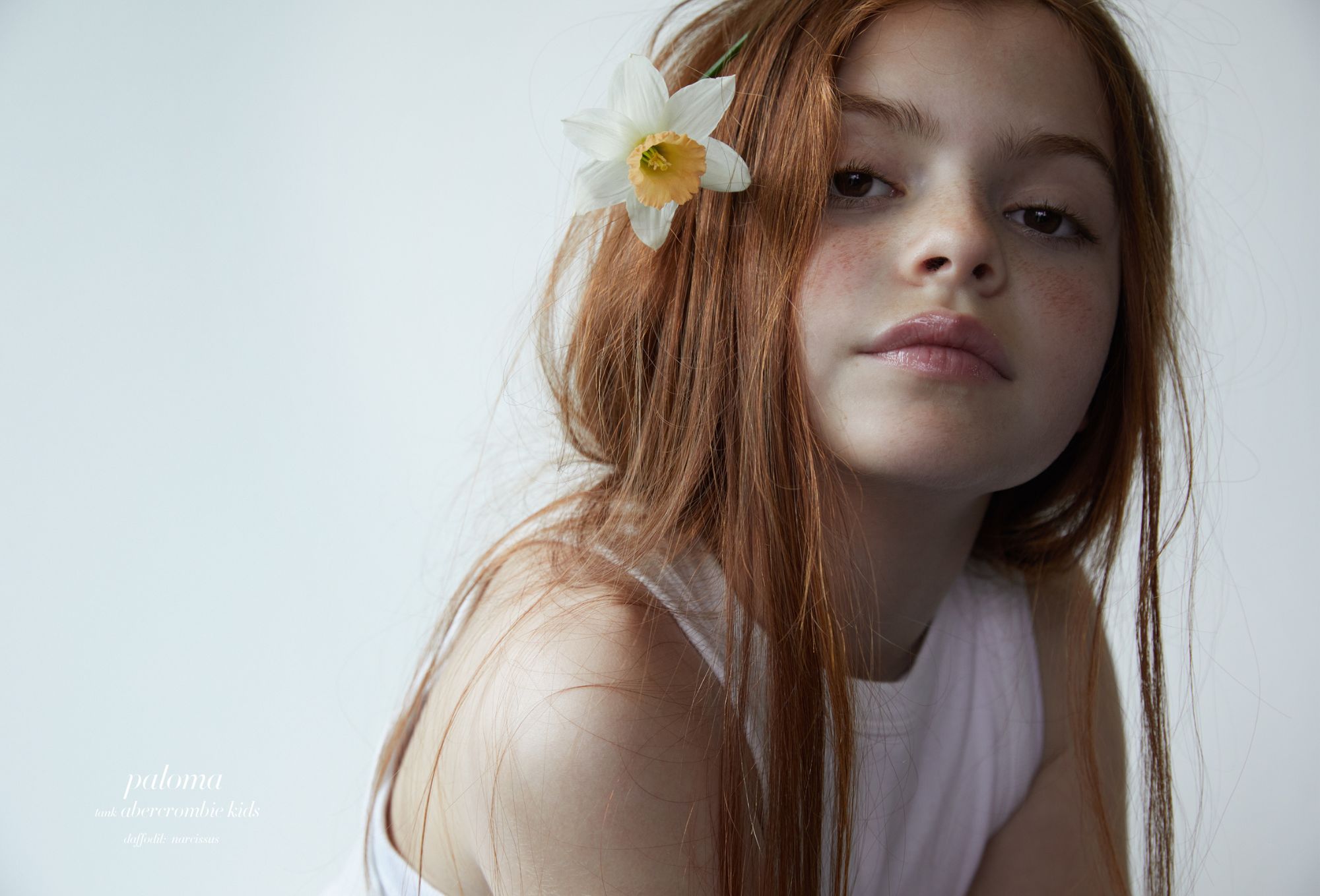Ritratto ravvicinato di bambina dai lunghi capelli rossi e lentiggini, narciso bianco tra i capelli, top bianco, su sfondo neutro; editoriale moda “Moon Garden” di Cleo Sullivan.