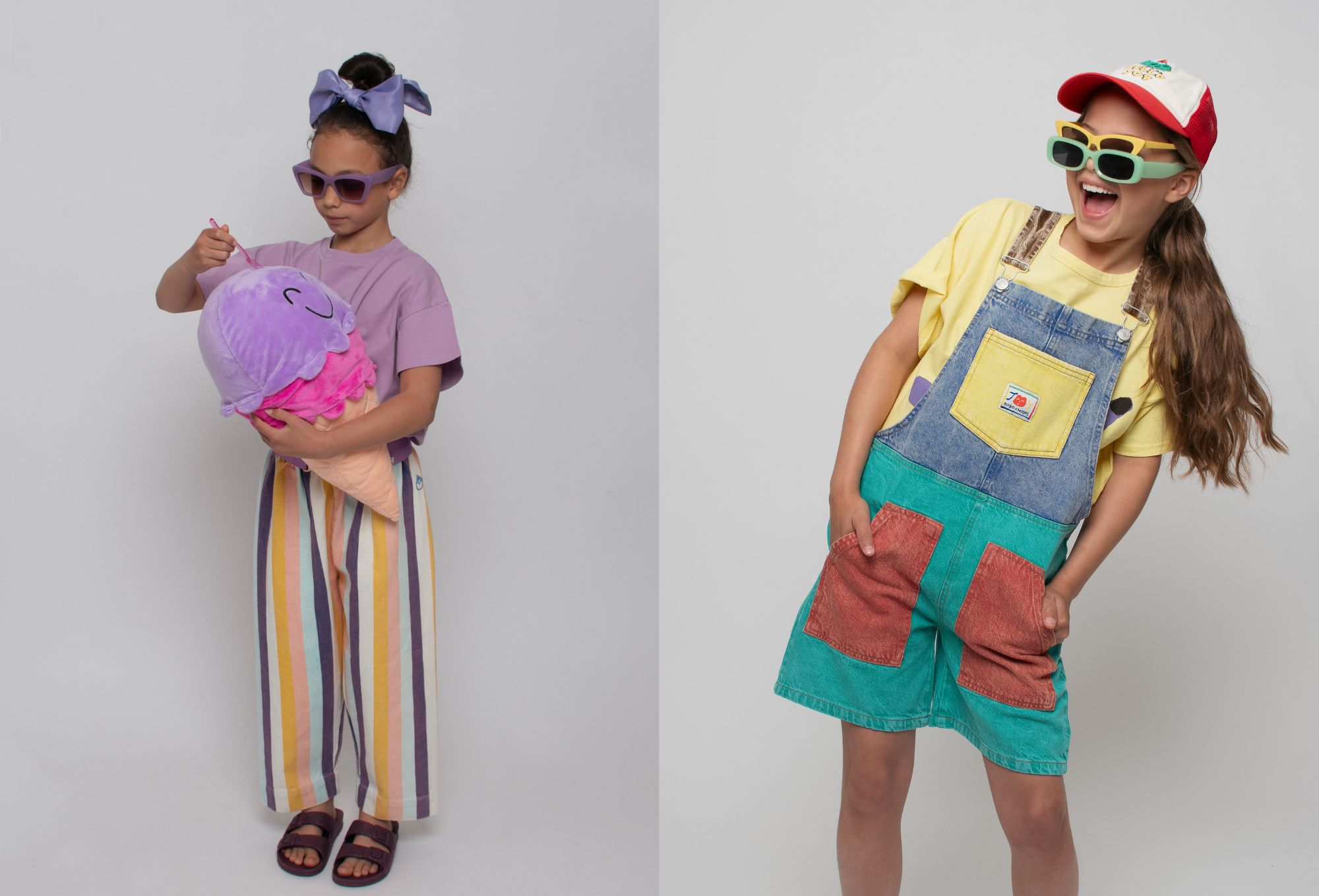 On the left, a girl in a lavender top and rainbow-striped pants pretends to scoop from a giant plush ice cream cone. On the right, another girl laughs in colorful patchwork denim overalls, layered over a yellow tee and topped with playful double sunglasses and a red cap.