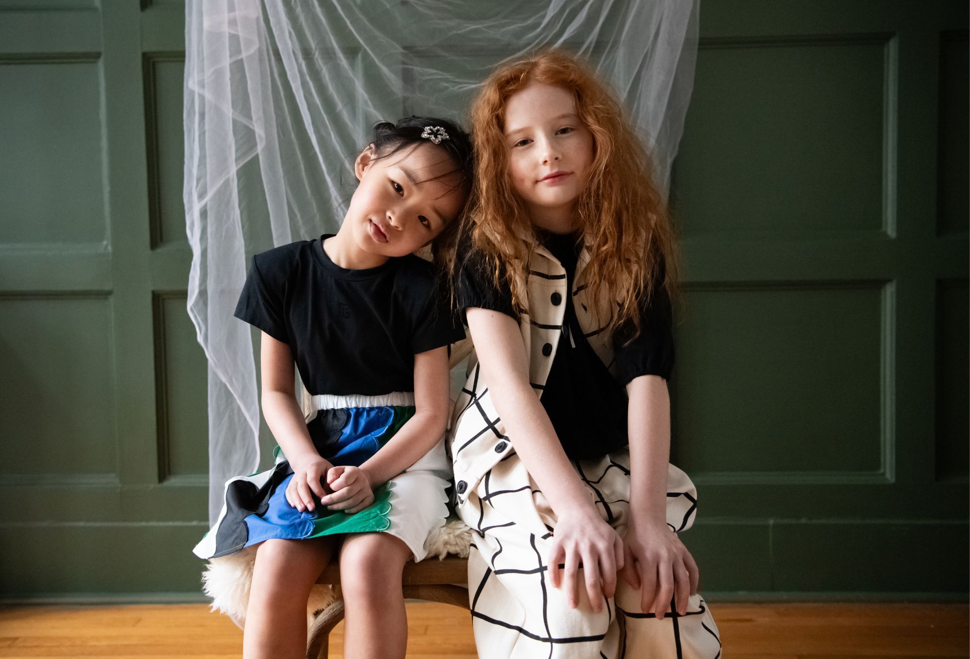 Two young girls sit closely together on a wooden chair with a soft white cushion, set against a green paneled wall and draped white tulle. The girl on the left leans her head affectionately on the other’s shoulder, wearing a black top and a color-blocked skirt in blue, green, and white. The girl on the right has long red hair and wears a black top paired with a cream outfit featuring bold black grid lines and large buttons. Both gaze softly at the camera. Photo by Sabrina Helas.