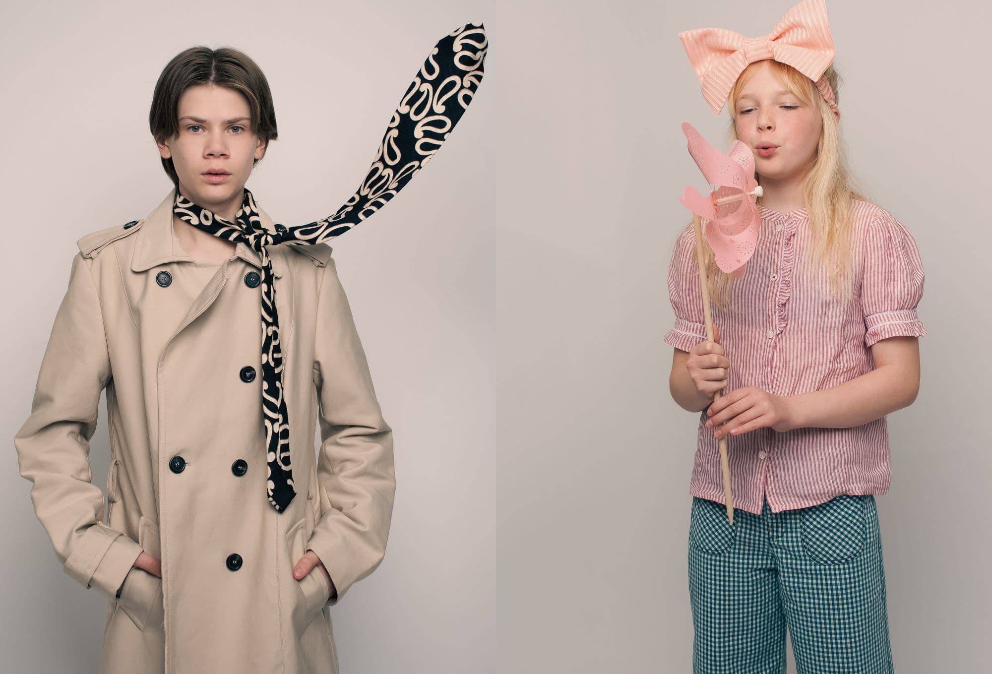 A moody boy in a beige trench coat with a patterned scarf flying mid-air; next to him, a girl in a pink striped blouse and oversized bow blows on a pastel pink pinwheel.