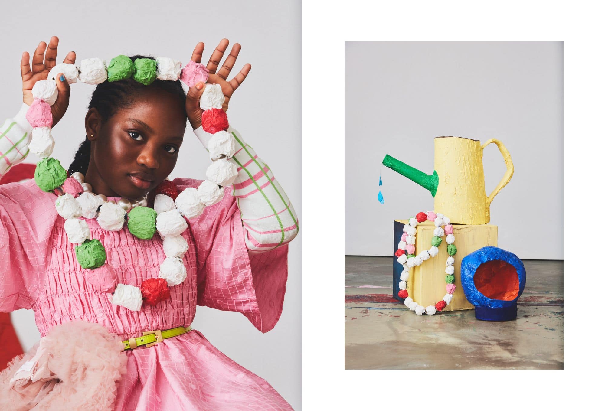 Children’s fashion editorial blending wearable art with playful props. On the left, a girl poses with a chunky necklace made of colorful paper-mâché beads in white, green, red, and pink tones, holding it up like a frame. She wears a vibrant pink smocked dress with puffed sleeves, a plaid-patterned undershirt, and a yellow belt with soft tulle details at the waist. On the right, a still life features the same oversized bead necklace draped over a yellow watering can, with a bold green spout and blue teardrop details. A sculptural blue-and-red bowl adds to the handcrafted, imaginative set.