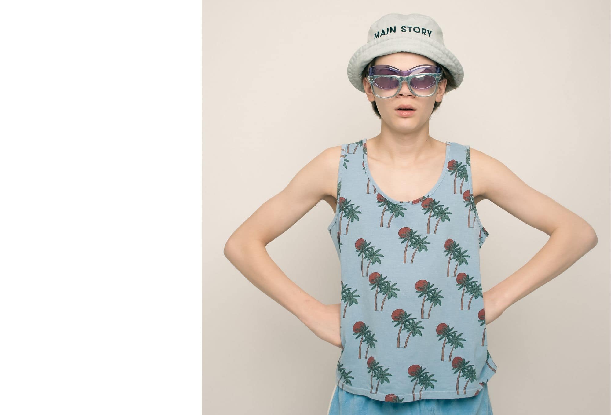 A cool teen wearing a palm tree tank top, double sunglasses, and a bucket hat with “Main Story” logo poses with hands on hips.