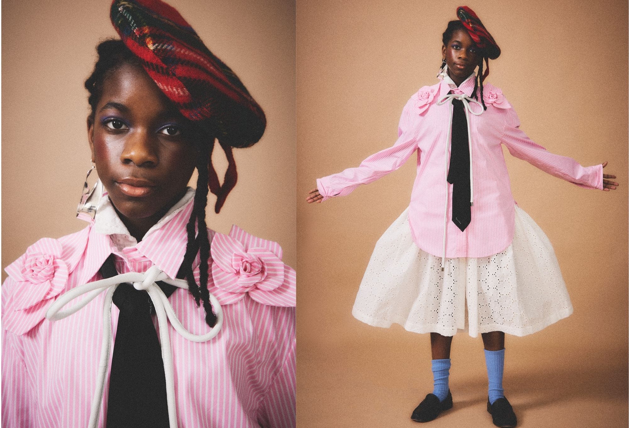 Fashion editorial by Molly Magnuson featuring a young model in a whimsical and avant-garde outfit. The look includes a pink and white striped shirt adorned with oversized fabric rosettes on the shoulders, a black necktie, and a dramatic white eyelet skirt with voluminous structure. The model wears a red plaid beret tilted to the side, a large sculptural earring, and light blue socks with black shoes. Set against a neutral background, the styling plays with proportions and textures, blending romantic details with school-uniform-inspired elements.