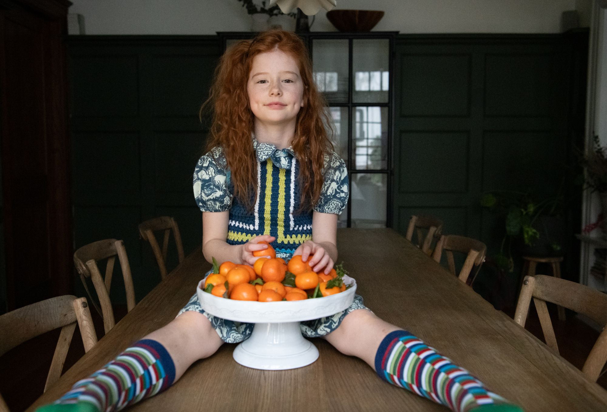 A young girl with long red hair sits playfully at the center of a wooden dining table with her legs stretched out, wearing a colorful crochet dress with a blue and yellow pattern over a puff-sleeve shirt featuring animal prints. In front of her is a white pedestal bowl filled with bright orange tangerines. She smiles softly, framed by striped knee-high socks and a cozy interior setting with wooden chairs and a green paneled wall in the background. Photo by Sabrina Helas.