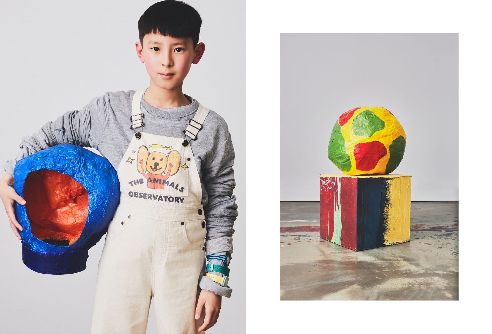 Artful children’s fashion editorial with a bold graphic style. On the left, a boy wears a grey sweatshirt layered under off-white overalls with a playful illustration of a cartoon dog and the words “THE ANIMALS OBSERVATORY.” He holds a large, hollow paper-mâché object painted blue on the outside and orange inside. Colorful watches peek from under his rolled-up sleeve, adding a pop of detail. On the right, a yellow, red, and green paper-mâché globe sits atop a hand-painted cube in distressed primary colors. The background remains minimal to highlight the handcrafted and imaginative world of the shoot.