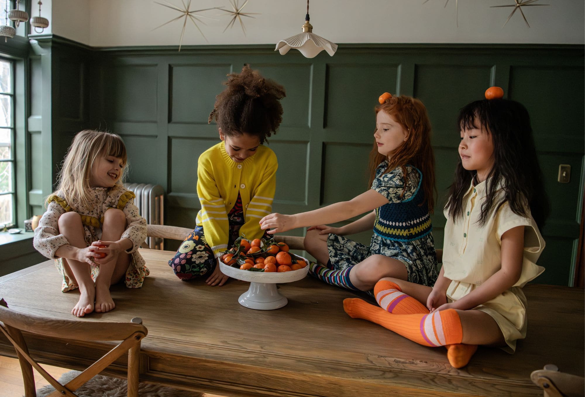 Alt text: Four young girls sit together on a large wooden table in a cozy, sunlit room with green paneled walls. In the center is a white pedestal bowl filled with vibrant tangerines. One girl, wearing a floral dress with a yellow collar, sits cross-legged and smiles while holding a fruit. Another girl in a yellow cardigan reaches toward the bowl, while the girl next to her wears a crochet vest and balances a tangerine on her head. The fourth girl, in a pale yellow romper with bright orange socks, also balances a tangerine on her head with a calm expression. Photo by Sabrina Helas.