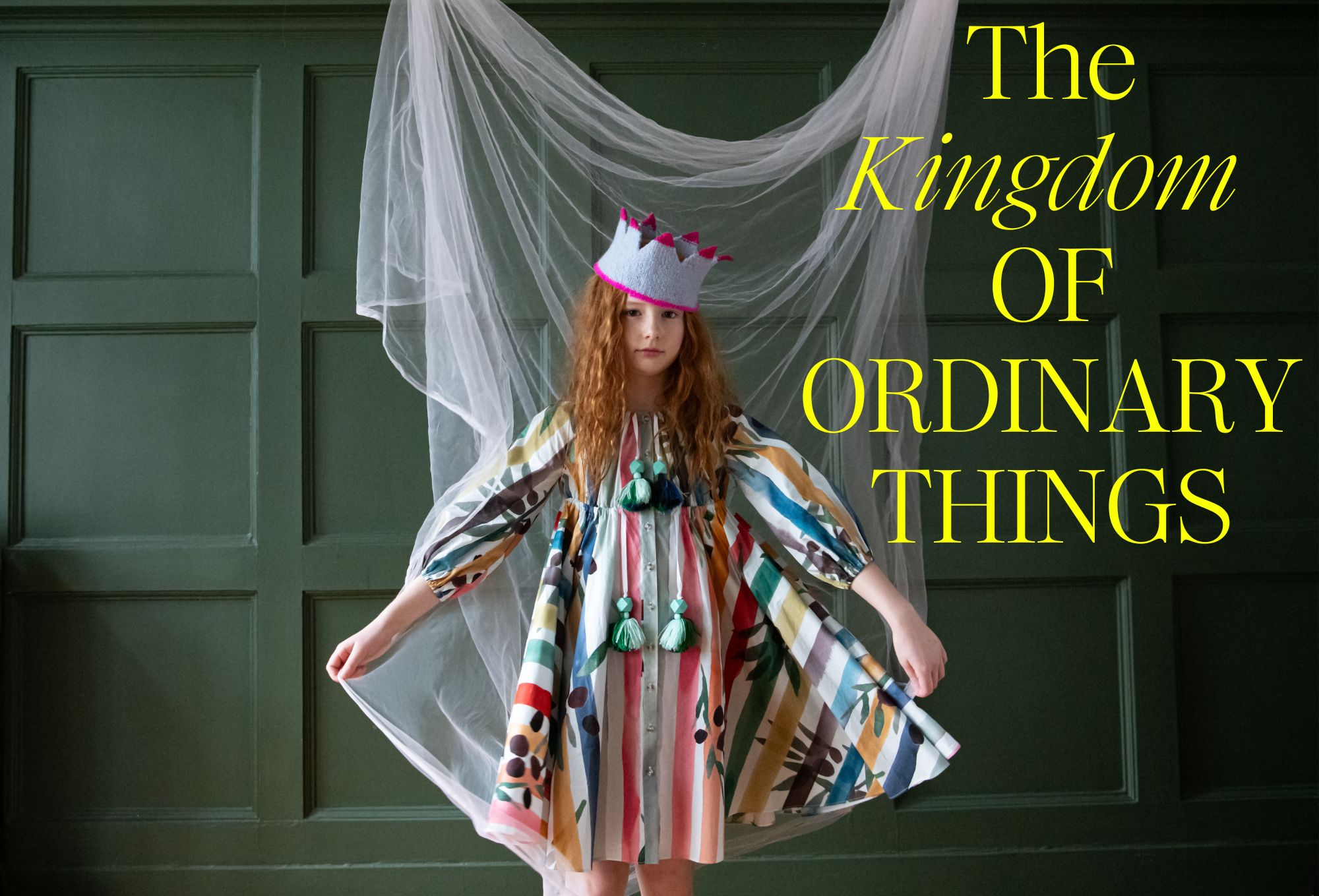 A young girl with long red hair stands in front of a dark green paneled wall, wearing a colorful, patterned dress with voluminous sleeves and green tassel details. She also wears a playful grey crown with pink accents. Behind her hangs a sheer white fabric, creating a dreamy, whimsical atmosphere. Text in yellow reads: “Kingdom of Ordinary Things.” Photo by Sabrina Helas.