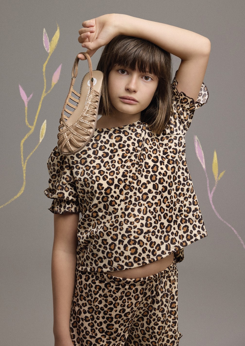 Girl wearing a matching leopard-print outfit holds a golden braided sandal above her head. The background is neutral gray with pastel-colored hand-drawn leaves, adding a playful touch to the composition.