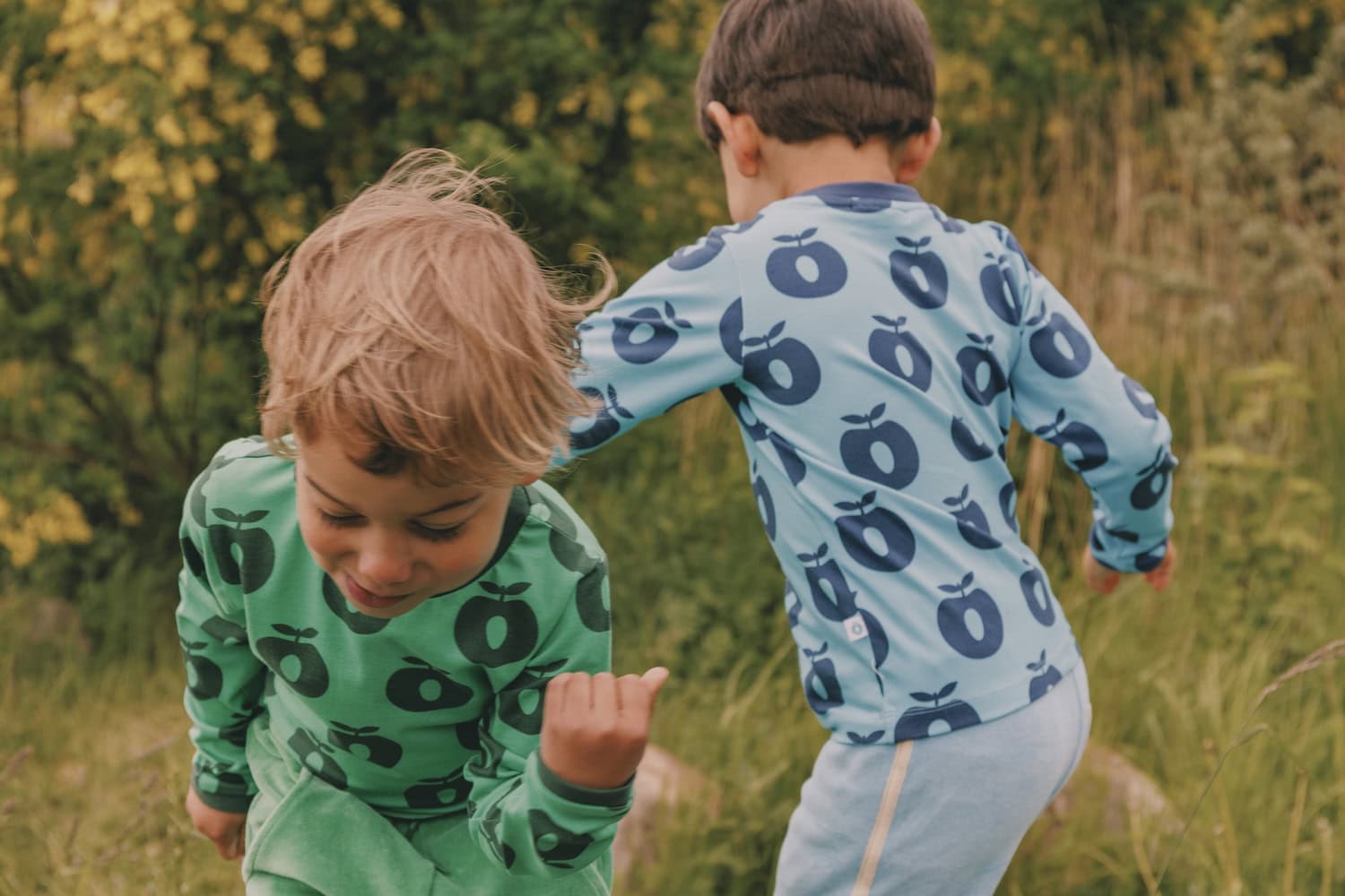 Two young children play joyfully outdoors in nature, wearing colorful long-sleeve tops from the Småfolk collection—one in green with dark green apple prints, the other in light blue with navy apple prints. The scene captures the carefree spirit of childhood among tall grass and yellow-flowered bushes.