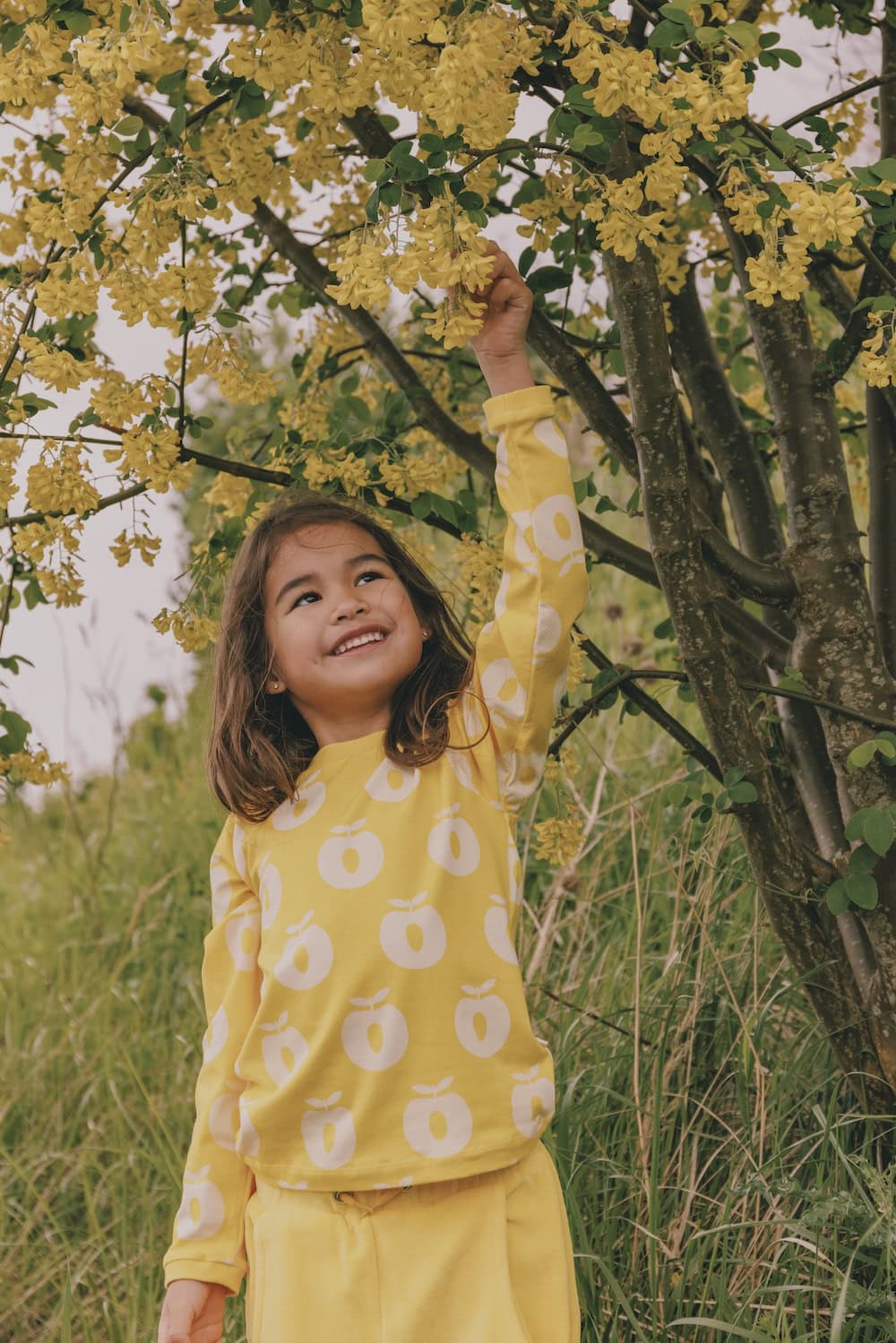 A smiling young girl stands under a blooming tree, reaching up to touch a cluster of bright yellow flowers. She wears a matching yellow outfit from the Småfolk collection, with a long-sleeve top featuring a white apple print. The scene radiates joy, nature, and playful harmony.