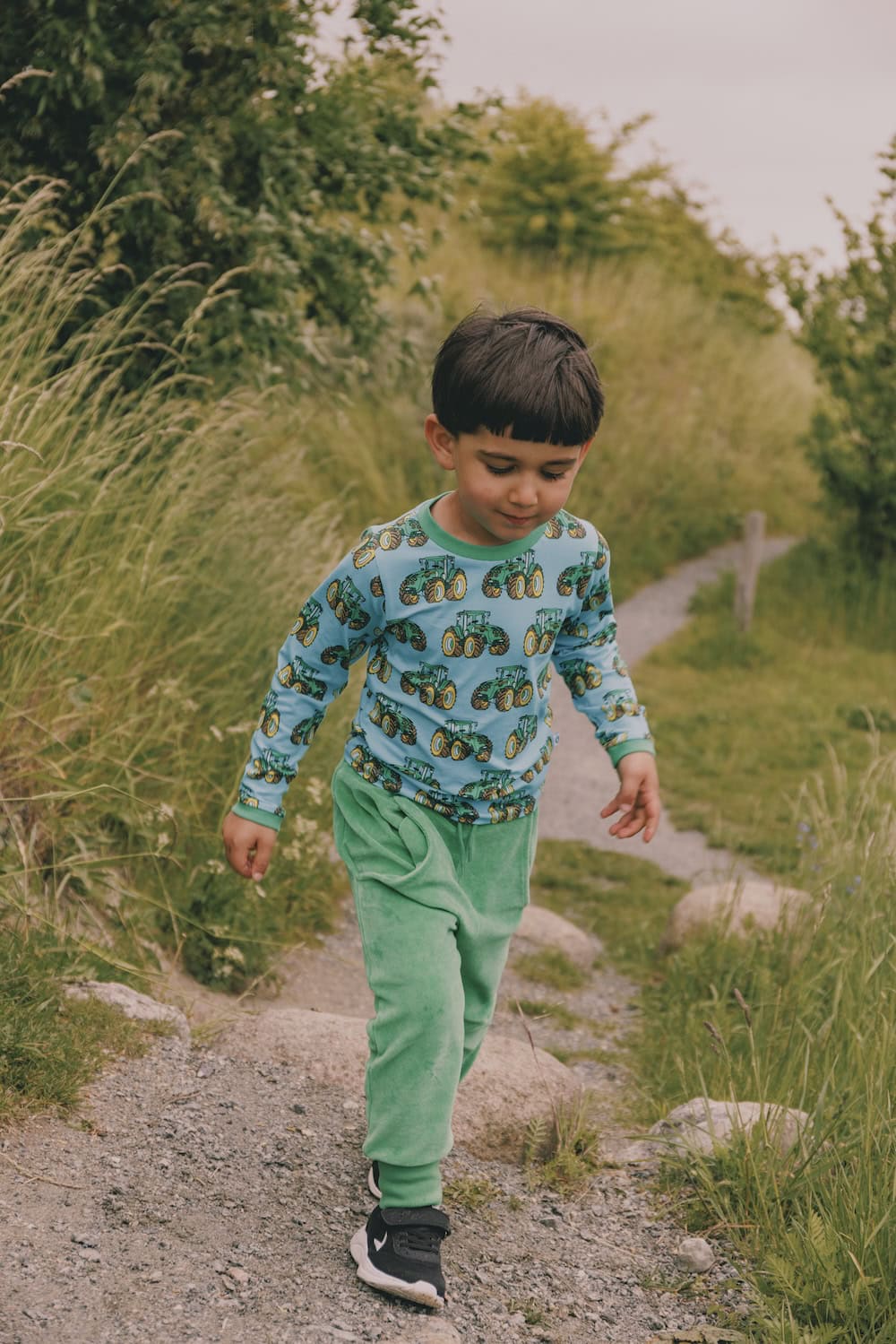 A young boy walks along a gravel path surrounded by tall grass, wearing a long-sleeve top with a green tractor print and green velour pants from the Småfolk collection. His expression is calm and focused, capturing a peaceful outdoor moment in nature.