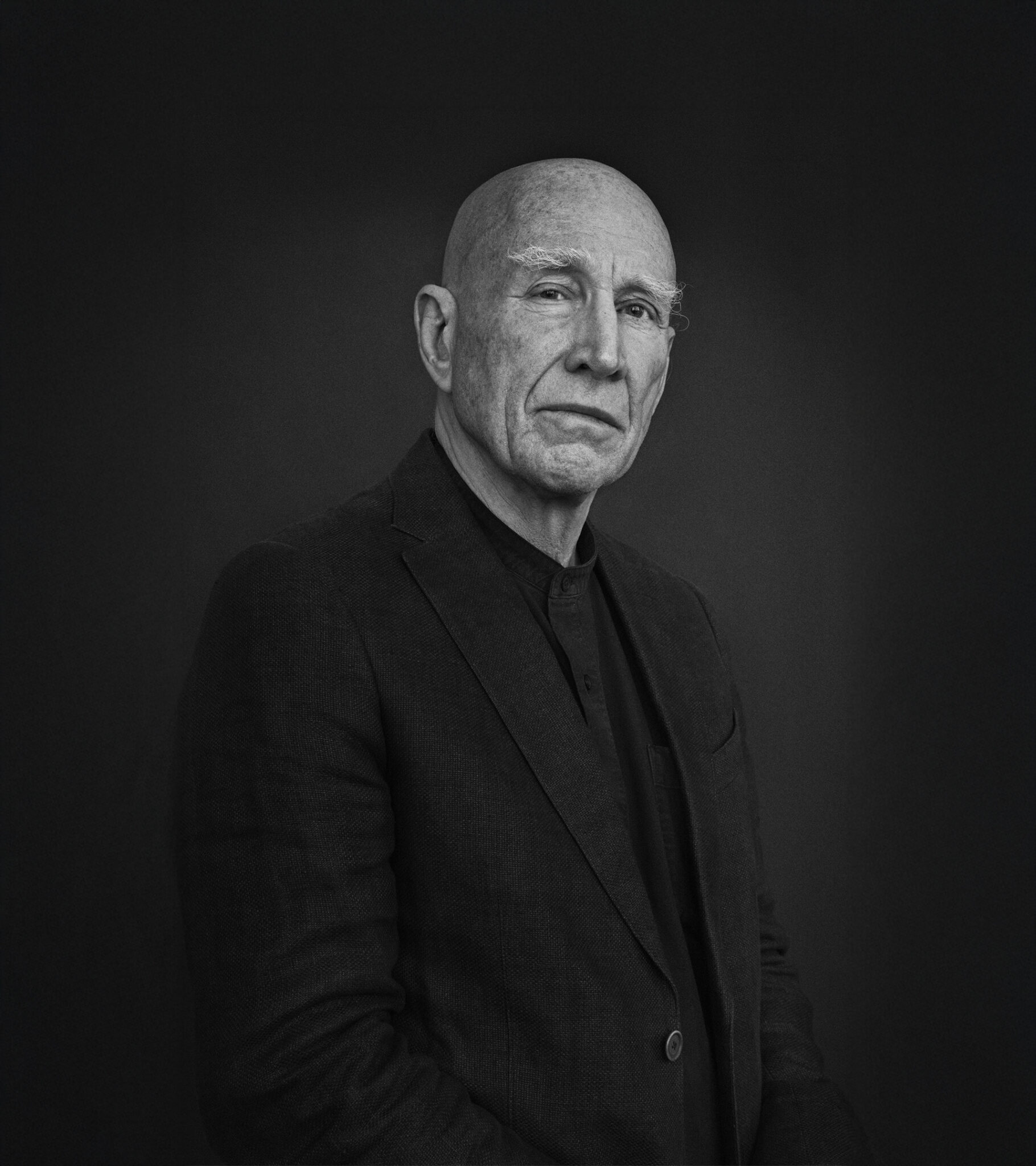 Black and white portrait of Sebastião Salgado, the renowned photographer, wearing a dark suit and looking solemnly at the camera against a plain dark background.