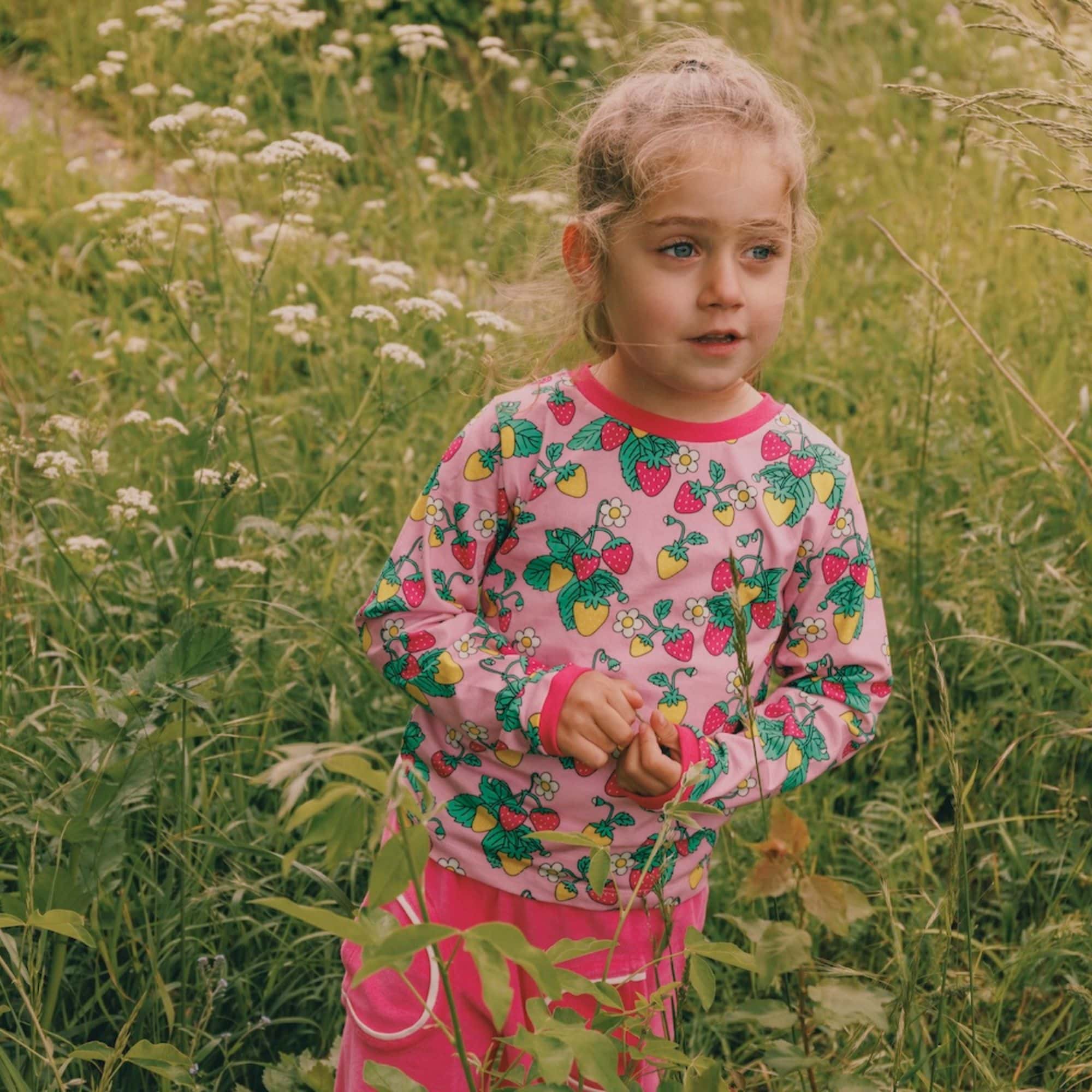 A little girl stands in a lush green meadow, surrounded by tall grass and white wildflowers. She wears a playful pink outfit from the Småfolk collection, featuring a long-sleeve top covered in a colorful strawberry print. Her thoughtful gaze and the soft breeze in her hair evoke a serene moment in nature.
