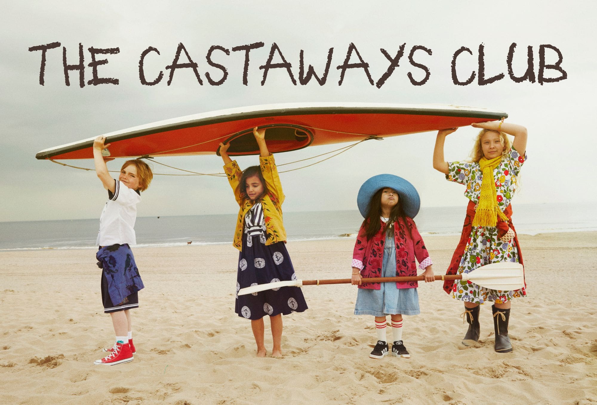 Four children stand on a sandy beach holding a red kayak above their heads. They wear colorful vintage-inspired outfits, including patterned dresses, sailor shorts, bold coats, and a wide-brimmed blue hat. The sea is calm in the background under a cloudy sky. The text