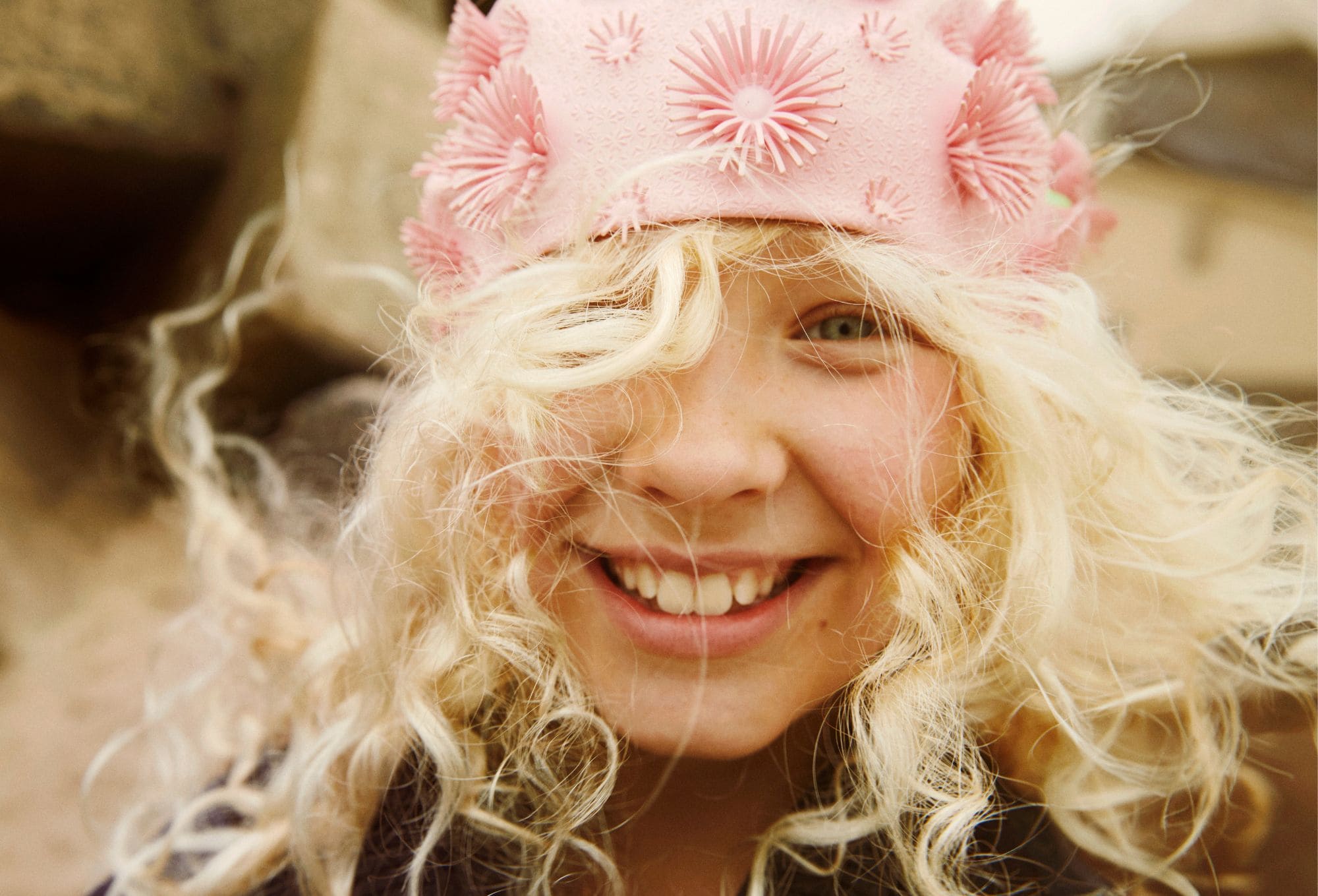 Close-up of a smiling child with long curly blonde hair, wearing a playful pink swim cap decorated with raised floral textures. The wind tousles their hair, and the background shows soft, sandy tones.