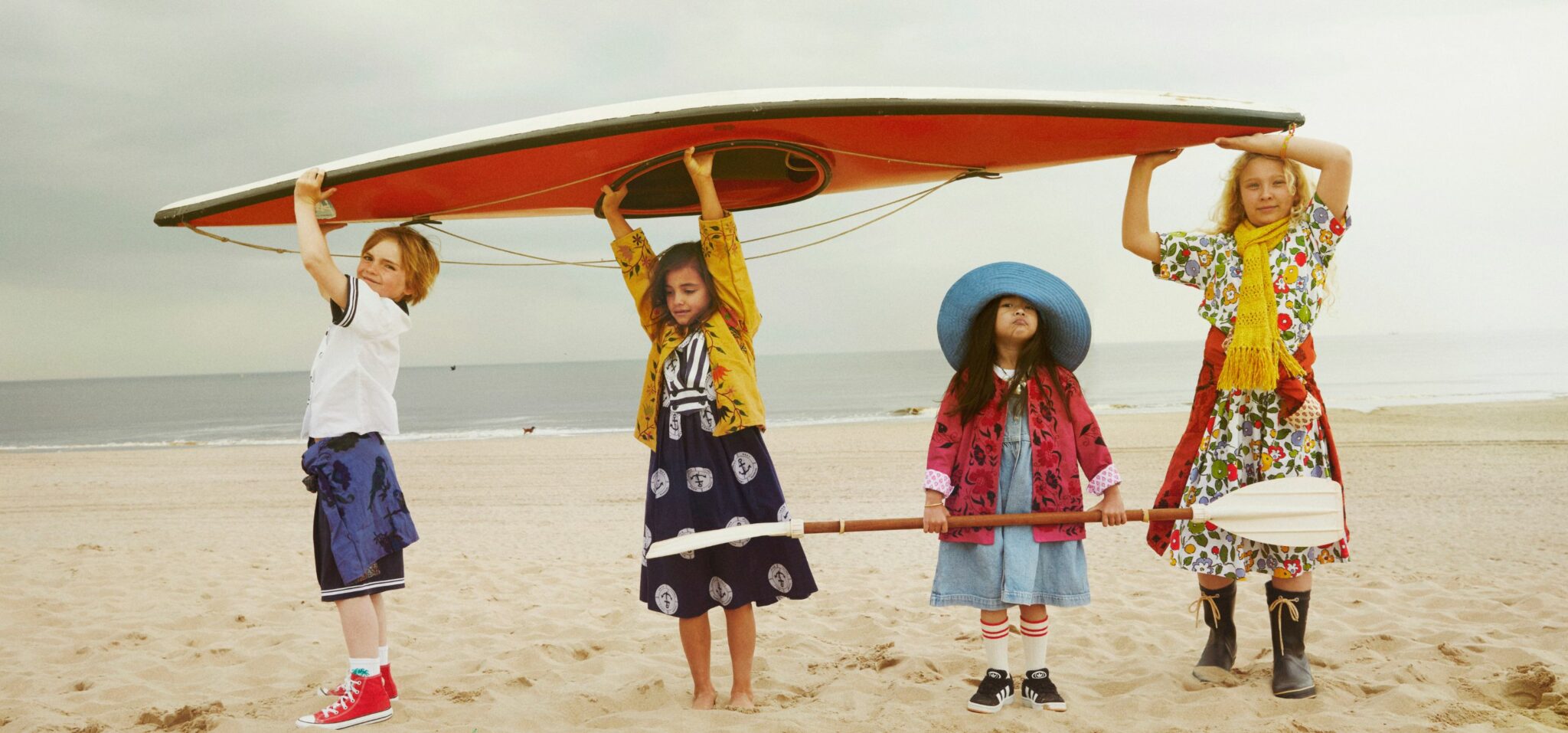 Four children stand on a sandy beach holding a red kayak above their heads. They wear colorful vintage-inspired outfits, including patterned dresses, sailor shorts, bold coats, and a wide-brimmed blue hat. The sea is calm in the background under a cloudy sky. The text
