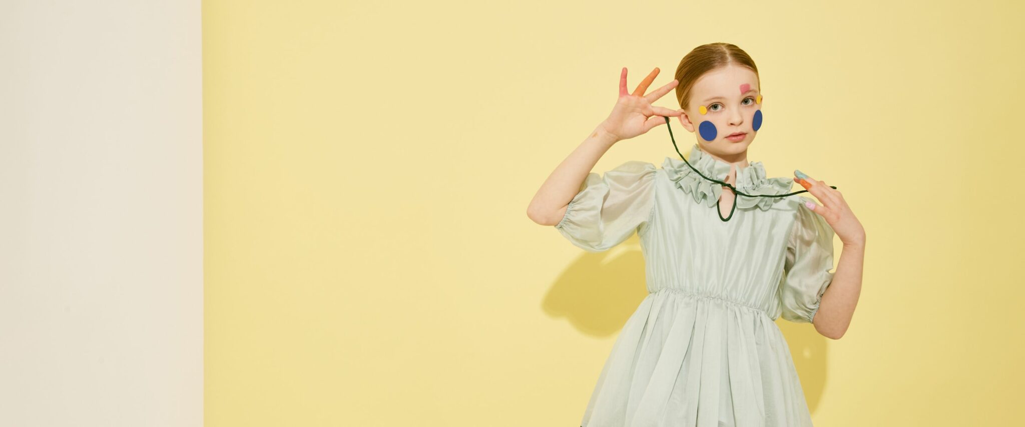 Una bambina posa davanti a uno sfondo giallo pallido indossando un abito verde menta con maniche a sbuffo e colletto arricciato, dalla collezione The Middle Daughter SS25. Il look è completato da adesivi colorati applicati sul viso e dita dipinte, creando un’atmosfera giocosa e teatrale.