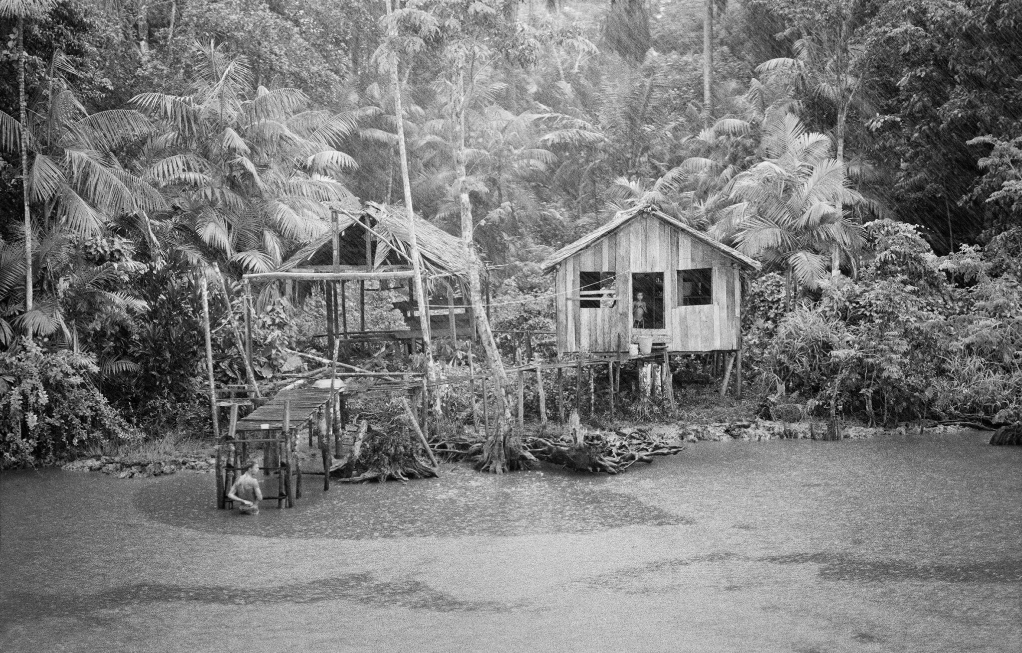 Black and white photograph from The Enchanted Ones by Stephanie Pommez, Kehrer Verlag, featured on Scimparello Magazine. Ribeirinho stilt house in the Brazilian Amazon surrounded by rainforest, with a person by the river under the rain.