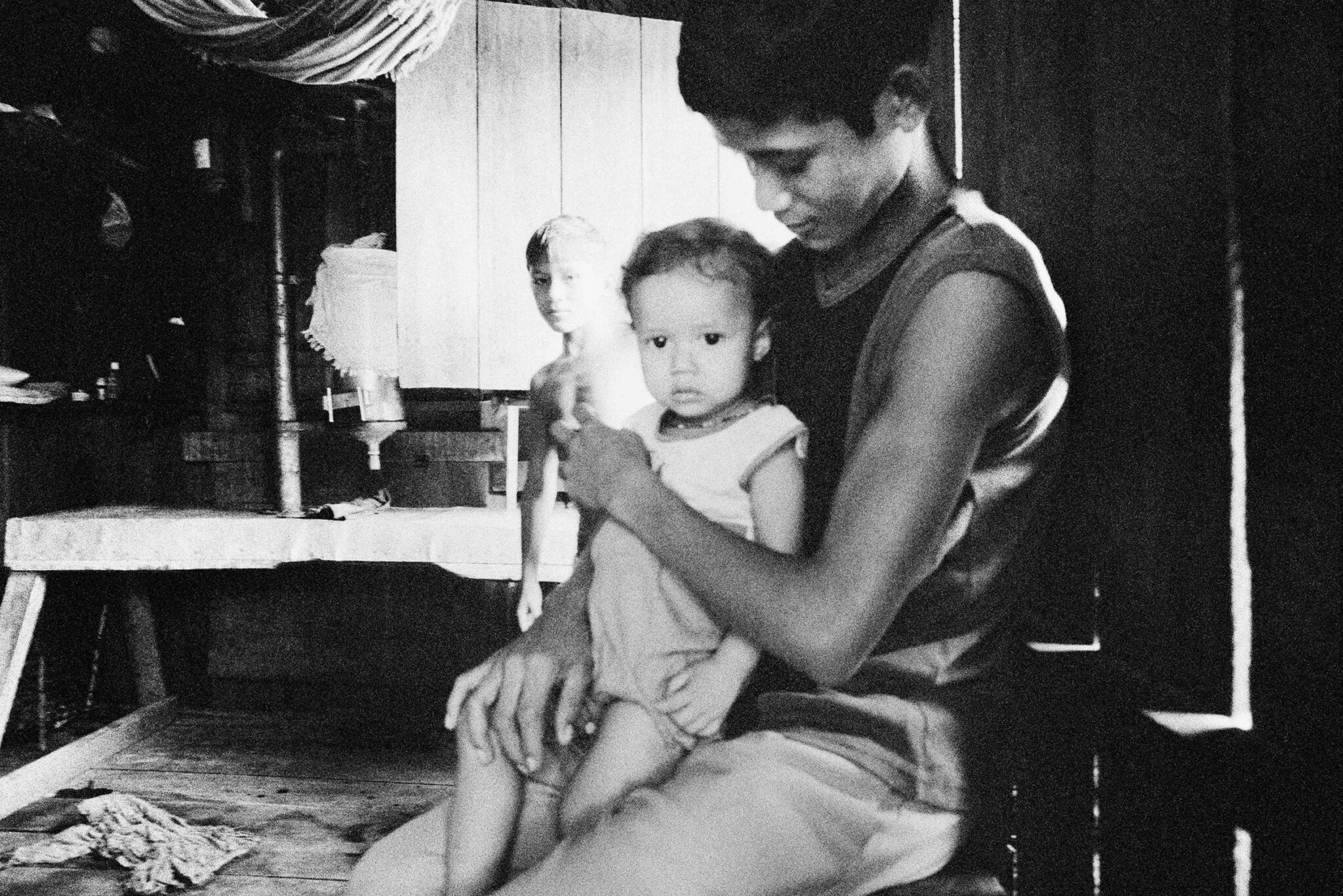 Black and white photographs from The Enchanted Ones by Stephanie Pommez, Kehrer Verlag, featured on Scimparello Magazine. Top: Ribeirinho midwife assisting childbirth in a poetic Amazonian scene. Bottom: young father holds a child inside a wooden house, another child in the background.