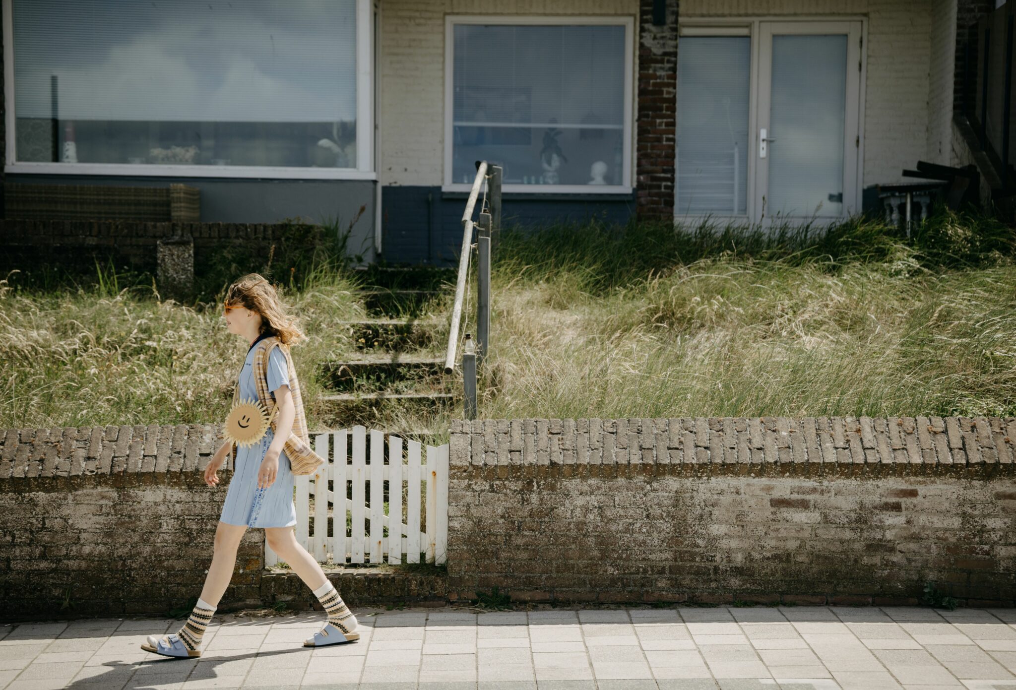 Teen fashion editorial Amsterdam Beach SS25, photographed by Arjaan Hamel for Scimparello Magazine. Girl walking along seaside houses wearing striped socks, sandals, blue dress, and sun-shaped bag.