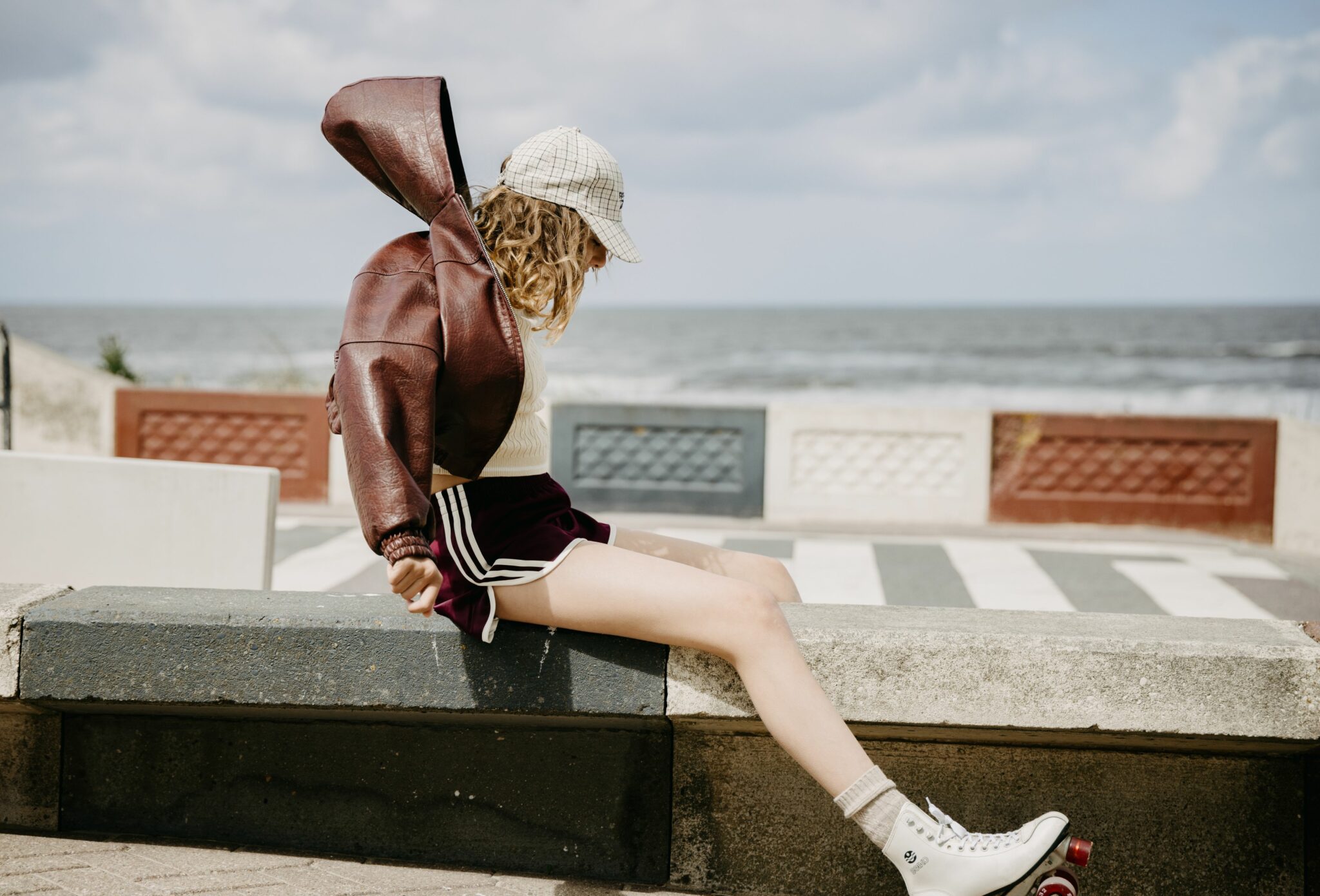 Teen girl in burgundy sports shorts, cream knit top, plaid cap, and white roller skates, sitting on a low wall by the seaside promenade in Amsterdam Beach, adjusting a brown leather hoodie jacket, photographed by Arjaan Hamel for Scimparello Magazine SS25 teen fashion editorial.