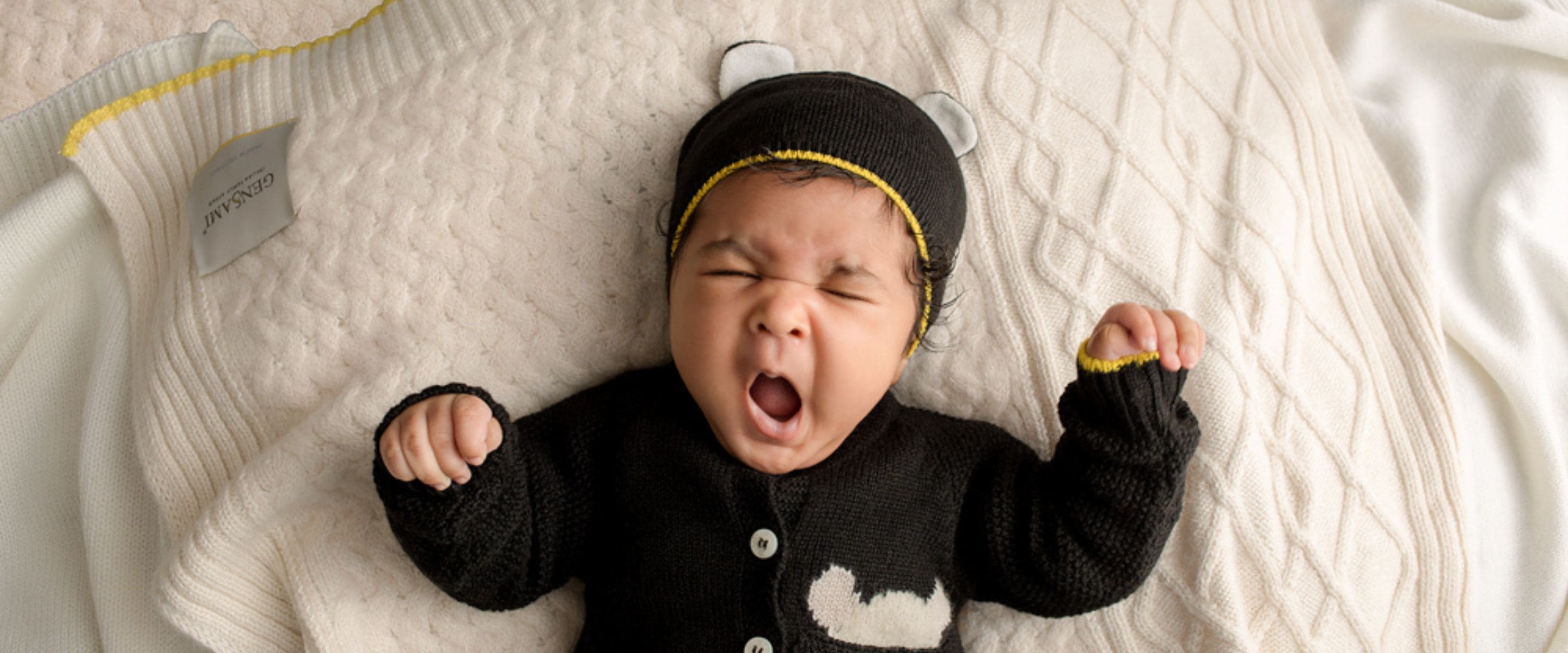 Baby in black knitted Gensami FW25 outfit with bear ear hat, lying on cream blanket.