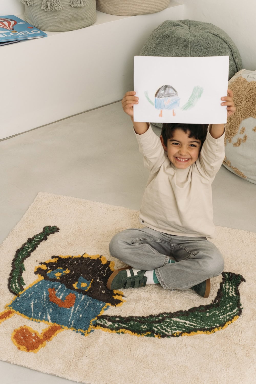 Smiling boy holding his drawing above his head while sitting on a handmade Lorena Canals rug featuring the same colorful design, from the Draw Your Own Rug collection.