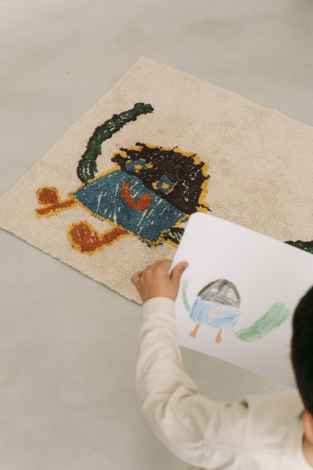 Child holding a drawing while looking at a handmade Lorena Canals rug featuring the same colorful design, part of the Draw Your Own Rug collection.