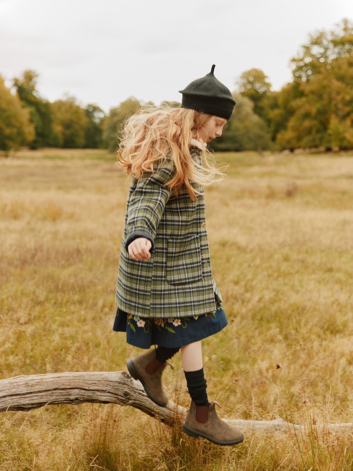 Girl in MOLO FW25 green plaid coat layered over a floral embroidered dress, styled with black knit hat and brown boots, walking on a fallen tree in a meadow.