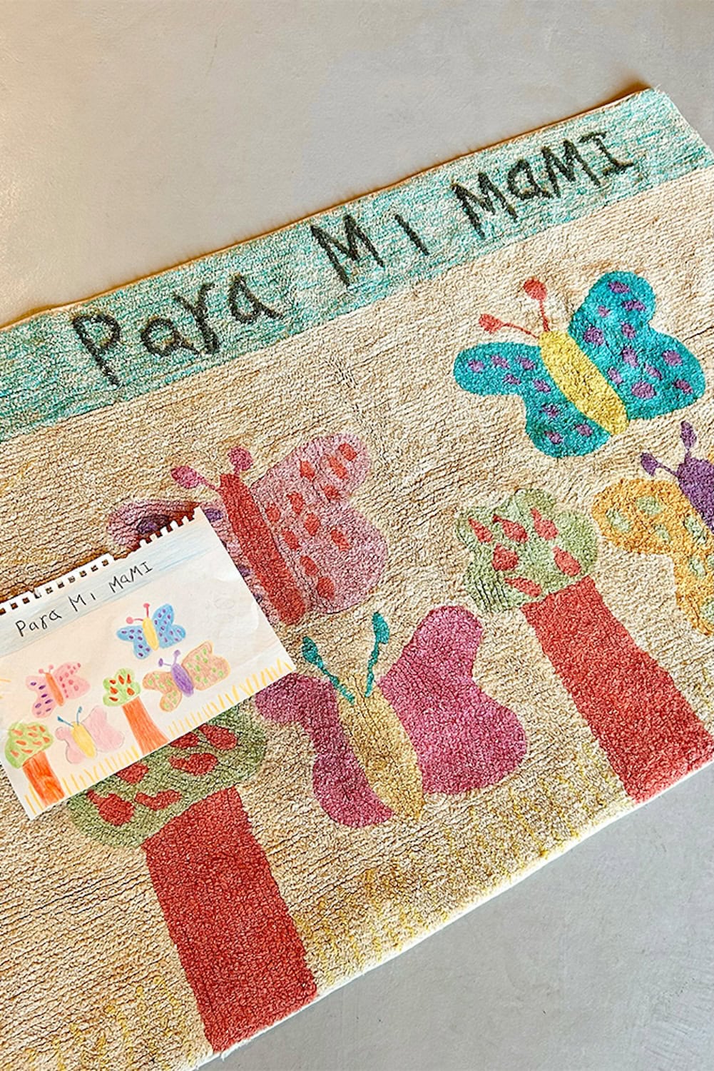 Handmade Lorena Canals rug with colorful butterflies and the words “Para Mi Mami,” shown alongside the original child’s drawing it was based on, part of the Draw Your Own Rug collection.