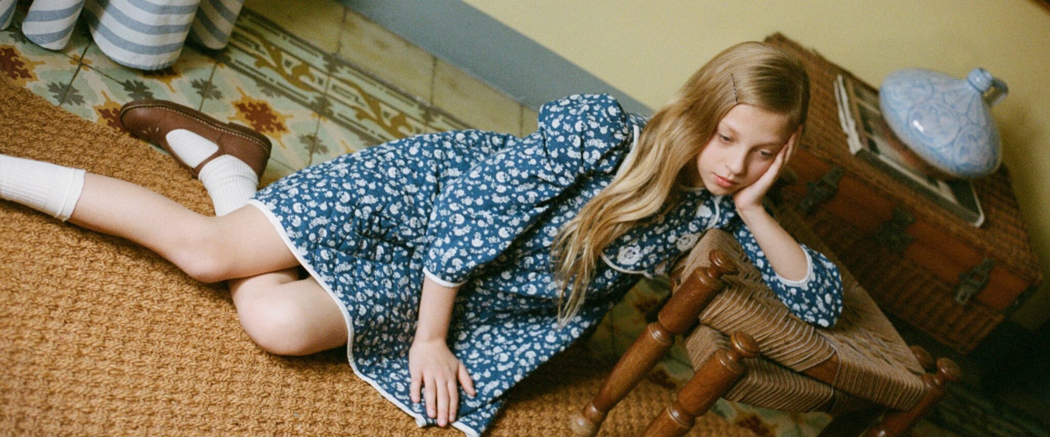 Girl lying on the floor in a vintage-inspired blue floral dress from Bebe Organic FW25, styled with white socks and brown shoes. Photo featured on Scimparello Magazine.