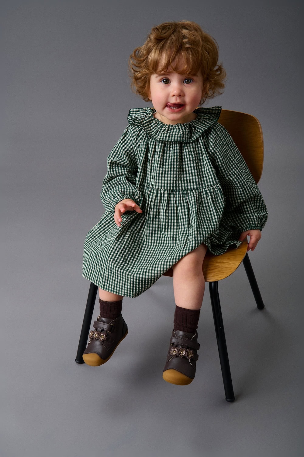 Toddler sitting on a wooden chair wearing a green gingham dress and brown leather shoes from BUNDGAARD FW25 kids shoe collection, Scimparello Magazine.