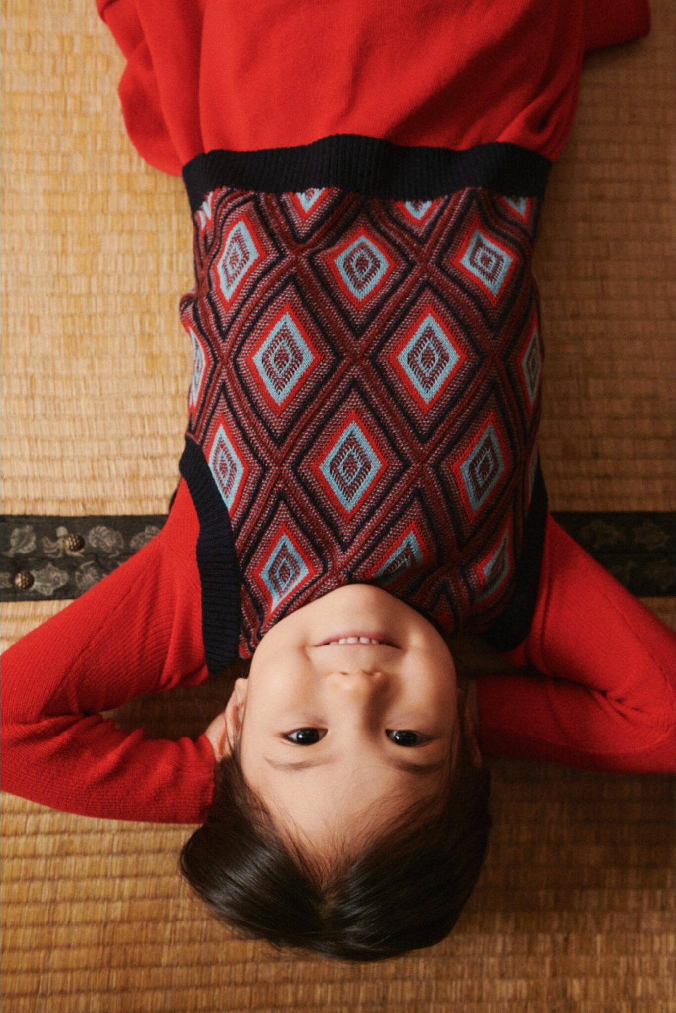 Child lying down smiling, wearing a red sweater layered with a geometric diamond-patterned knit vest in red, blue, and black – FUB Kids FW25.